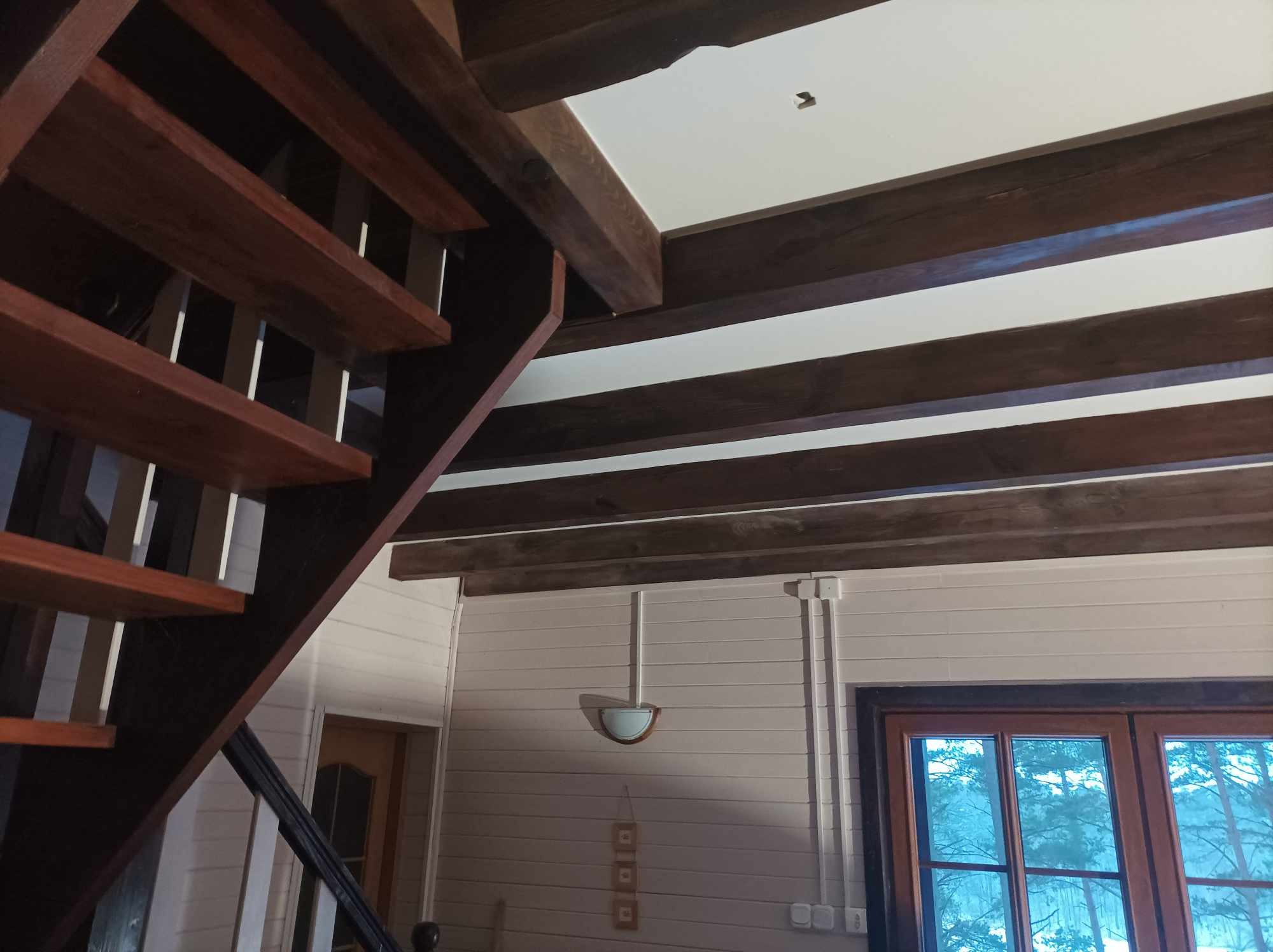 Wnętrze domu z drewnianymi schodami, belkami stropowymi i boazerią w jasnych odcieniach, widok na okno z widokiem na zewnątrz.