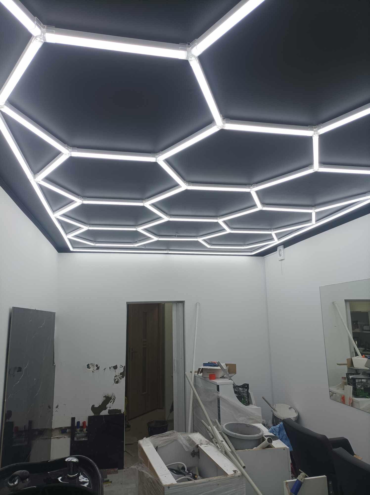 Sufit podwieszany z ciemnymi panelami w kształcie sześciokątów, oświetlony białymi, liniowymi lampami LED tworzącymi geometryczny wzór, widoczne elementy instalacji i niedokończone ściany...