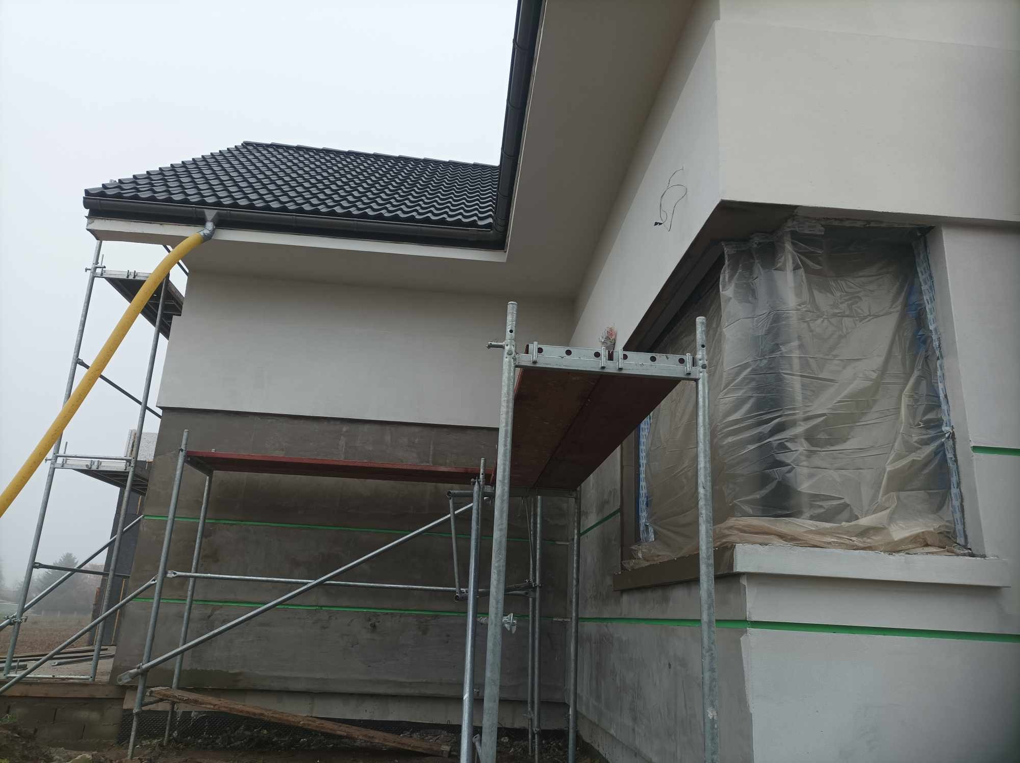 Elewacja domu w trakcie prac wykończeniowych: ocieplanie, nałożony tynk szary, okna zabezpieczone folią, rusztowanie, żółty przewód wentylacyjny