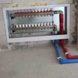 Rozdzielacz ogrzewania podłogowego zamontowany w ścianie, z widocznymi rurami doprowadzającymi wodę w kolorach czerwonym i niebieskim, w surowym stanie wykończenia.