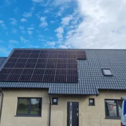 Nowoczesny dom jednorodzinny z szarym dachem pokrytym panelami fotowoltaicznymi, na tle błękitnego nieba z obłokami; widoczny fragment firmowego samochodu z napisem 'Pokrycia Dachowe'.