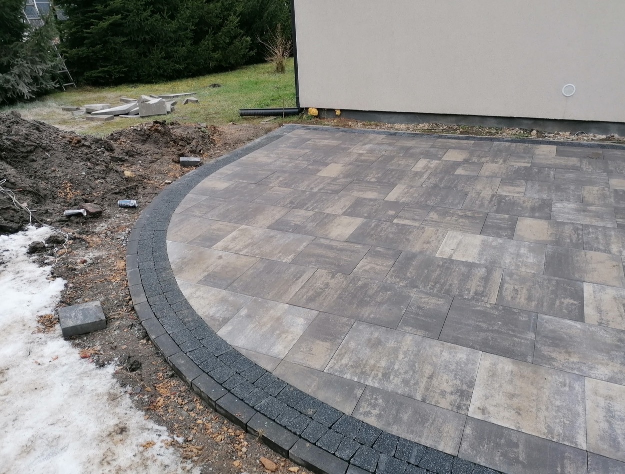 Ułożona kostka brukowa w odcieniach szarości i beżu, z ciemnoszarą obwódką, częściowo pokryta śniegiem przy krawędzi, z widoczną stertą ziemi i elementami kostki w tle.
