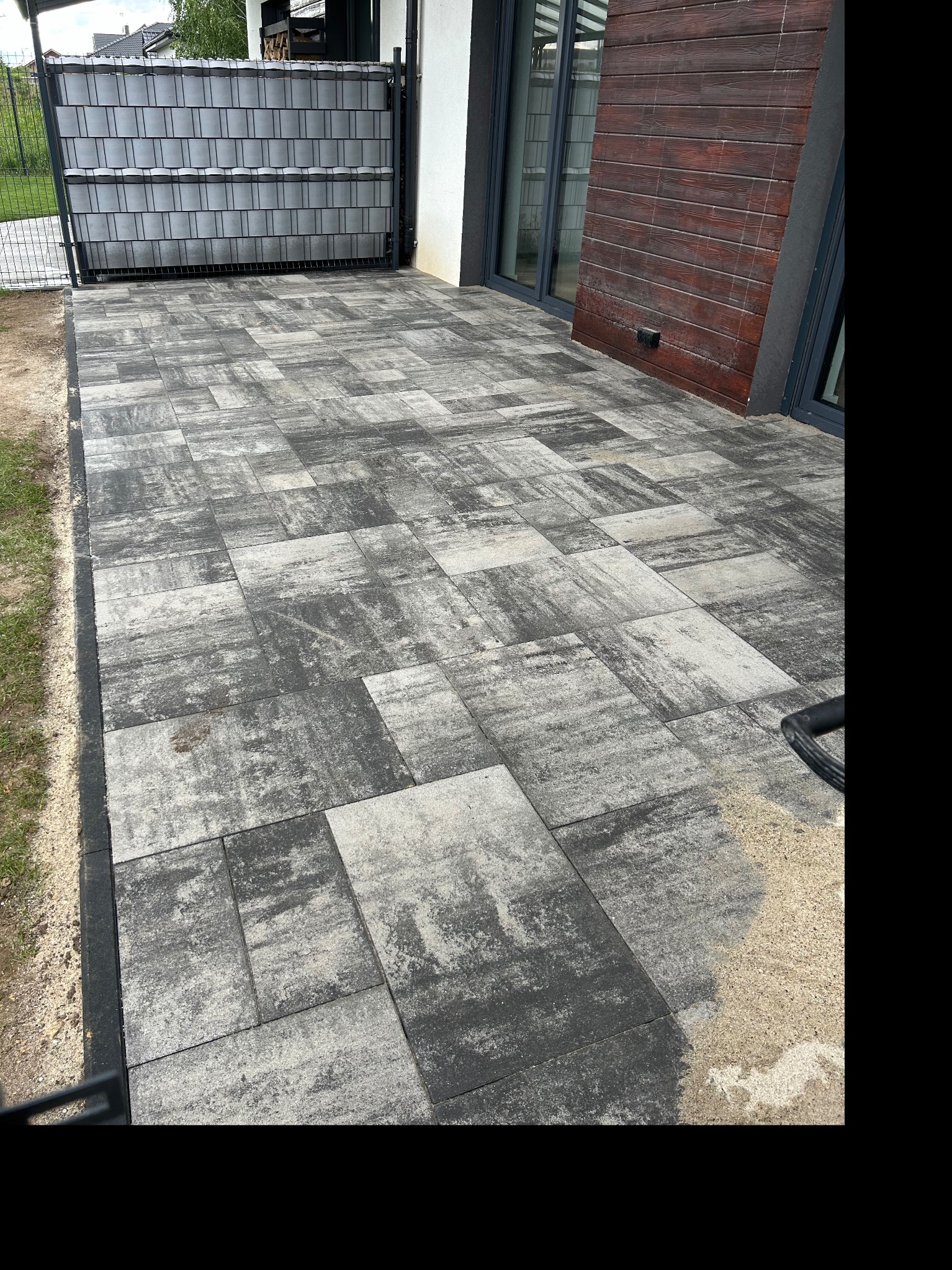 Ułożona szara kostka brukowa na posesji, widoczne obrzeża, fragment ogrodzenia panelowego i elewacja budynku z drewnopodobną okładziną.