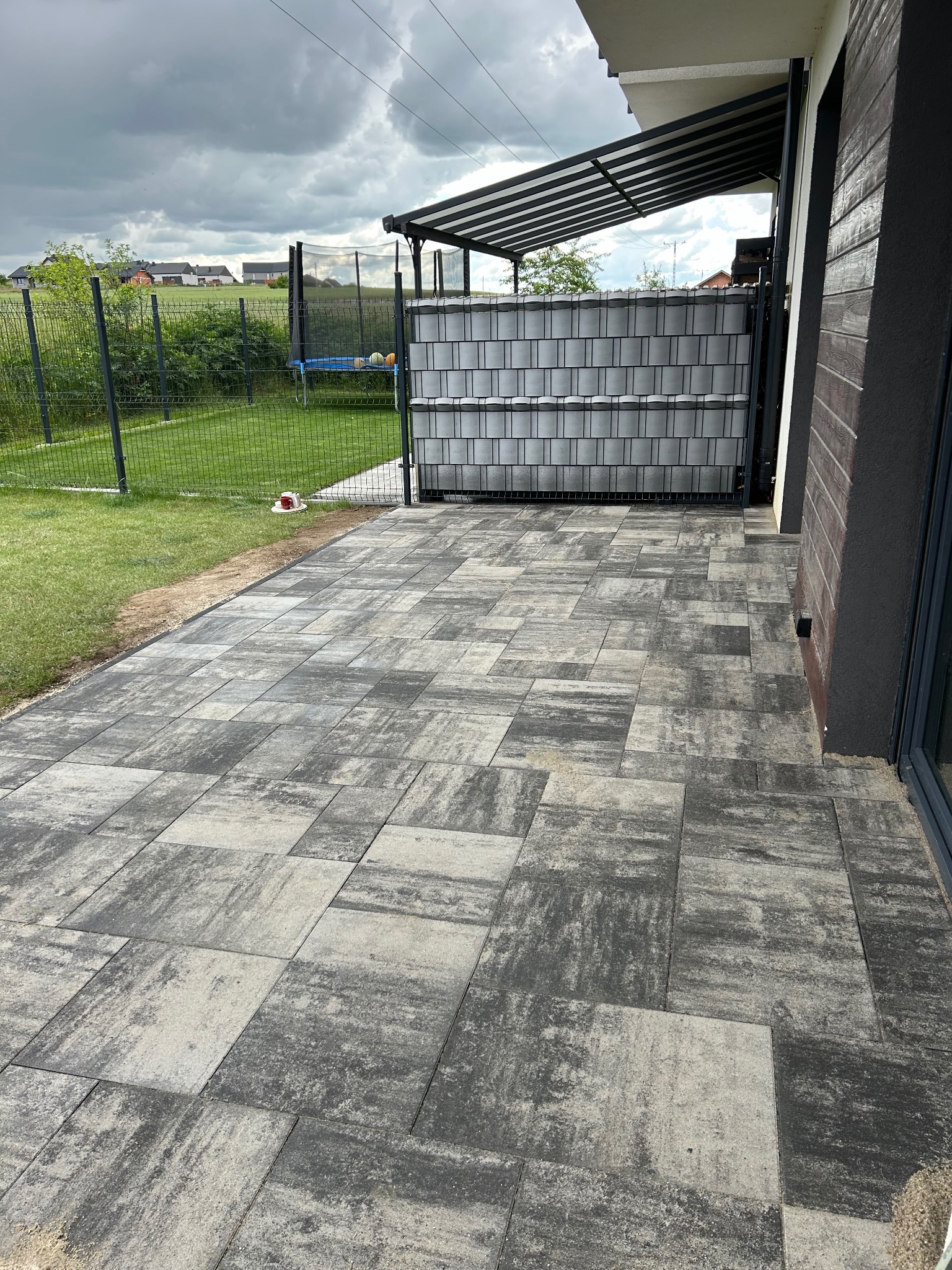 Nowo ułożony, szary chodnik z kostki brukowej o nieregularnym wzorze, prowadzący do bramy z panelami ogrodzeniowymi, widok na trawnik i domy w tle pod pochmurnym niebem.