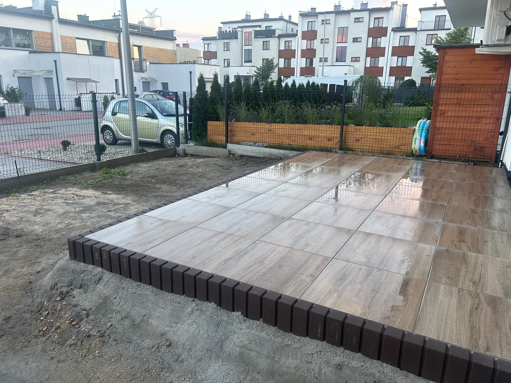 Wyłożony płytkami taras z obrzeżem z kostki, widok na domy w tle i fragment ogrodzenia, lekko mokra powierzchnia.