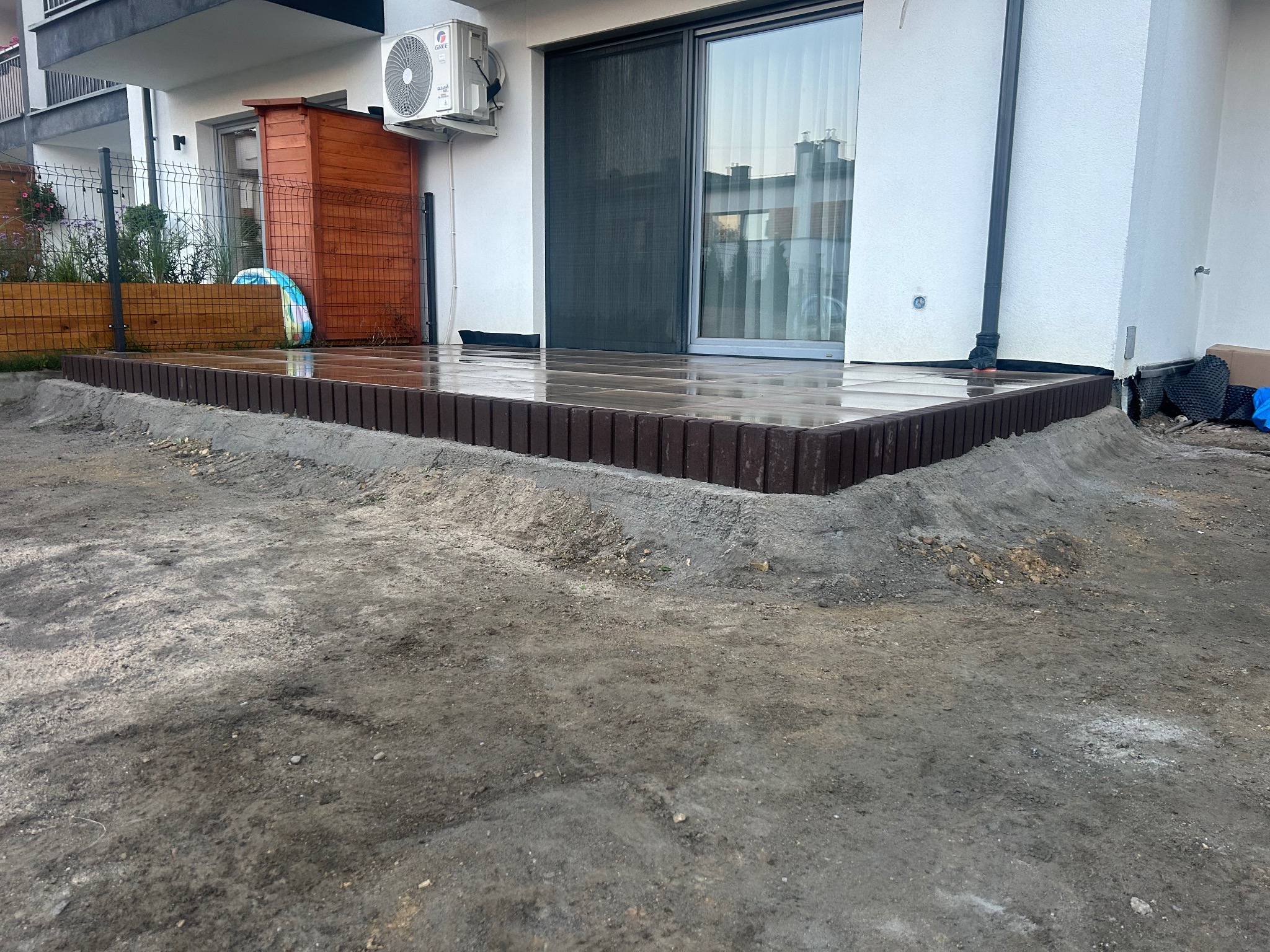 Taras z kompozytowych desek, obrzeża z ciemnobrązowej kostki, przygotowany teren wokół, widok na biały budynek z klimatyzatorem i przesuwnymi drzwiami tarasowymi.