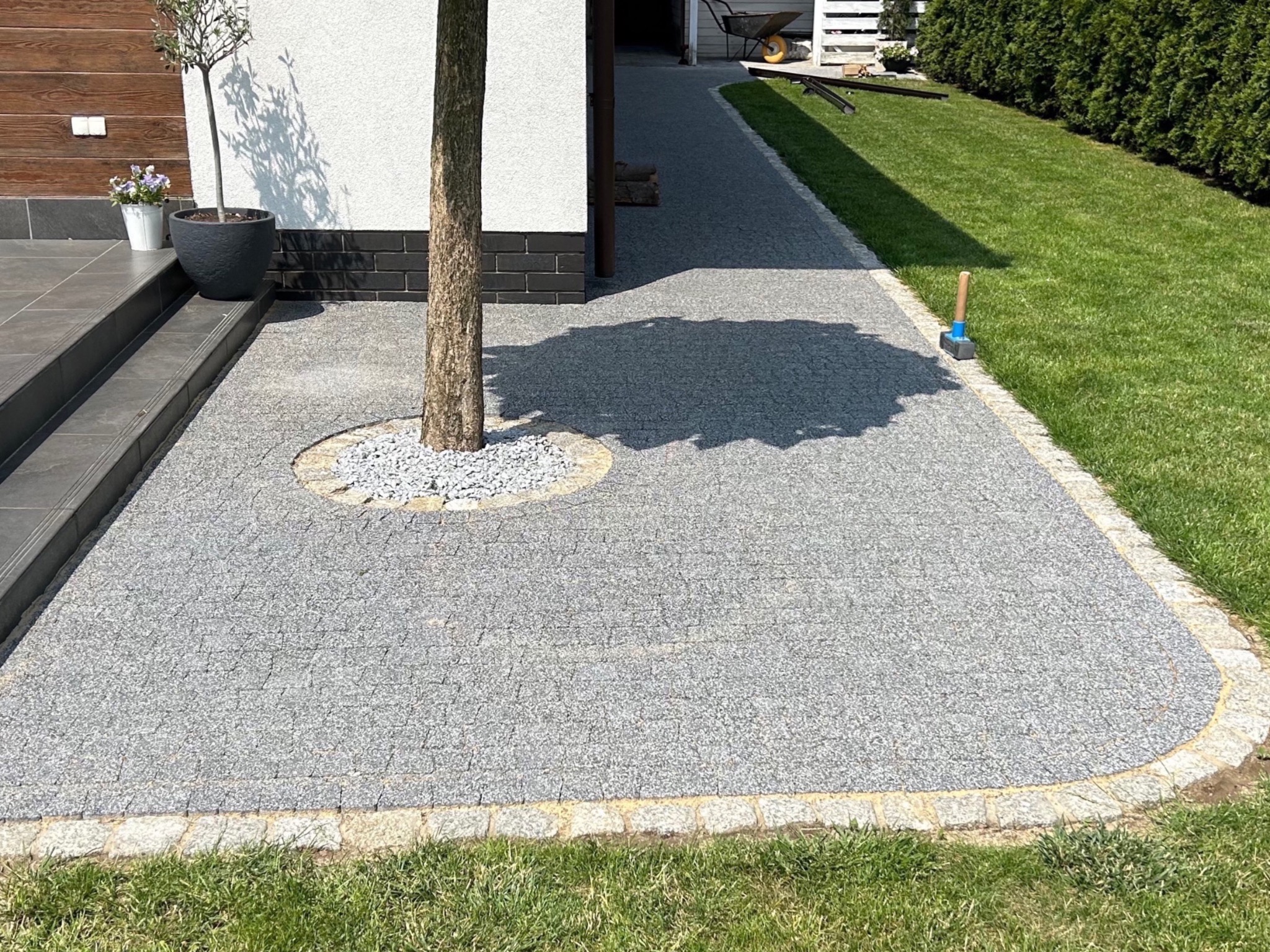 Szara nawierzchnia z drobnej kostki brukowej otaczająca drzewo, z obrzeżem z jasnego kamienia, prowadząca do domu z elementami drewnianej elewacji i zielonego trawnika