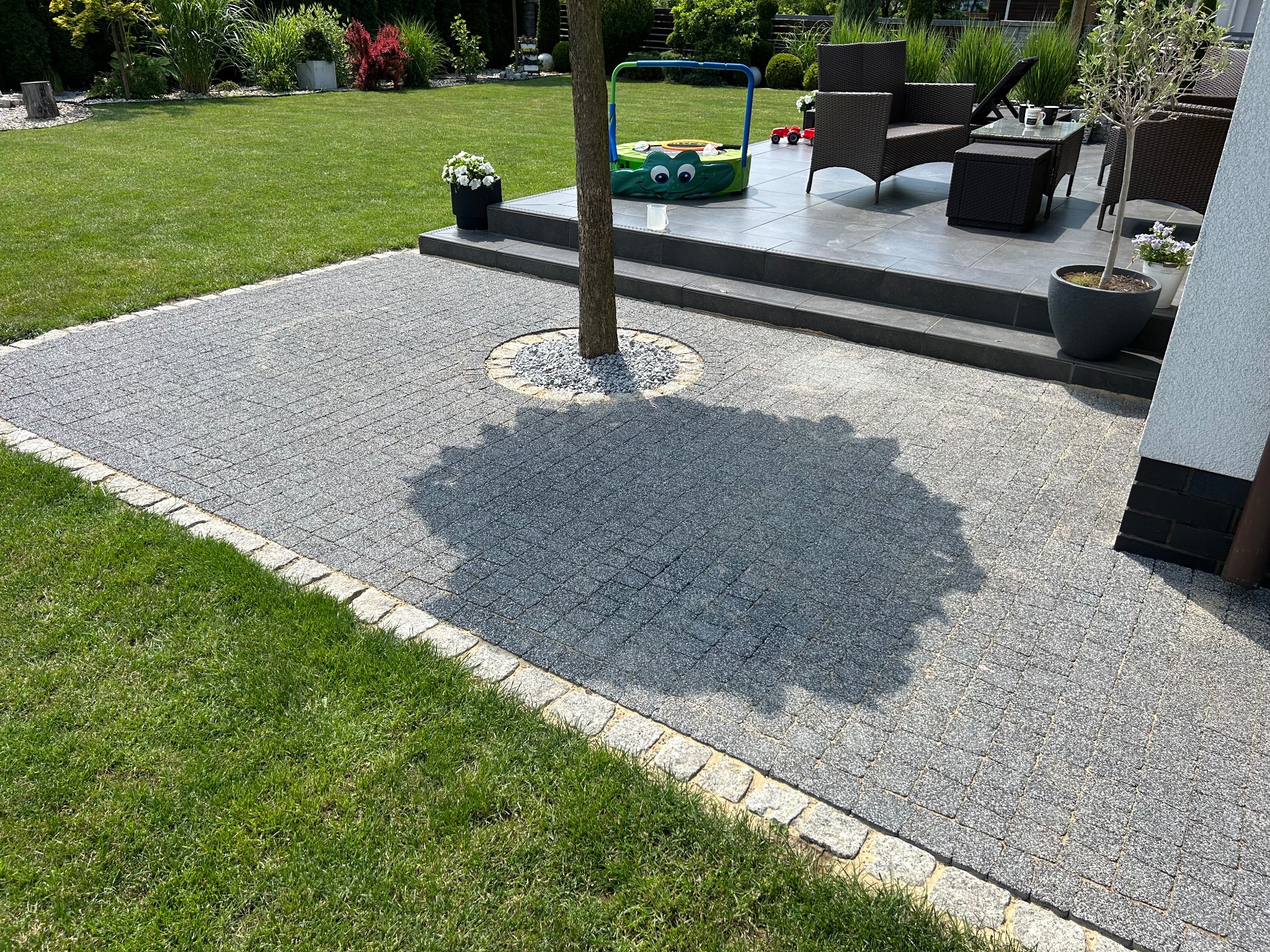 Szara kostka brukowa ułożona wokół drzewa z obrzeżem z jasnego kamienia, widok z góry na taras z meblami ogrodowymi i trawnik w słoneczny dzień.