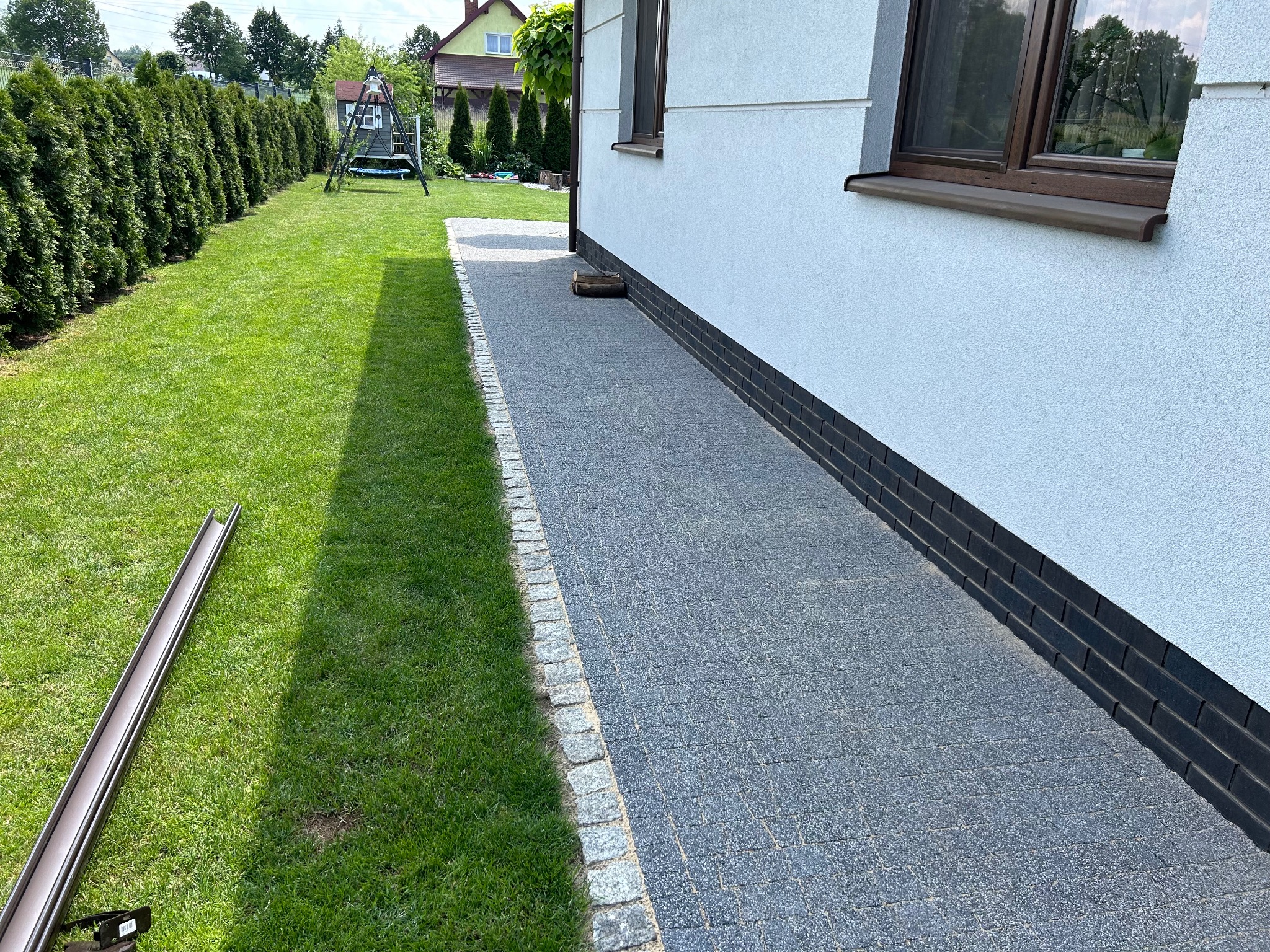 Ułożona ścieżka z szarej kostki brukowej obramowana jasnymi kamieniami, przylegająca do białego domu z ciemnymi oknami i otoczona zielonym trawnikiem, z widoczną rynną na trawie.