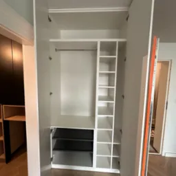 Wnętrze białej szafy wnękowej z drzwiami na zawiasach, widoczne półki, drążek i wysuwane kosze w kolorze czarnym.