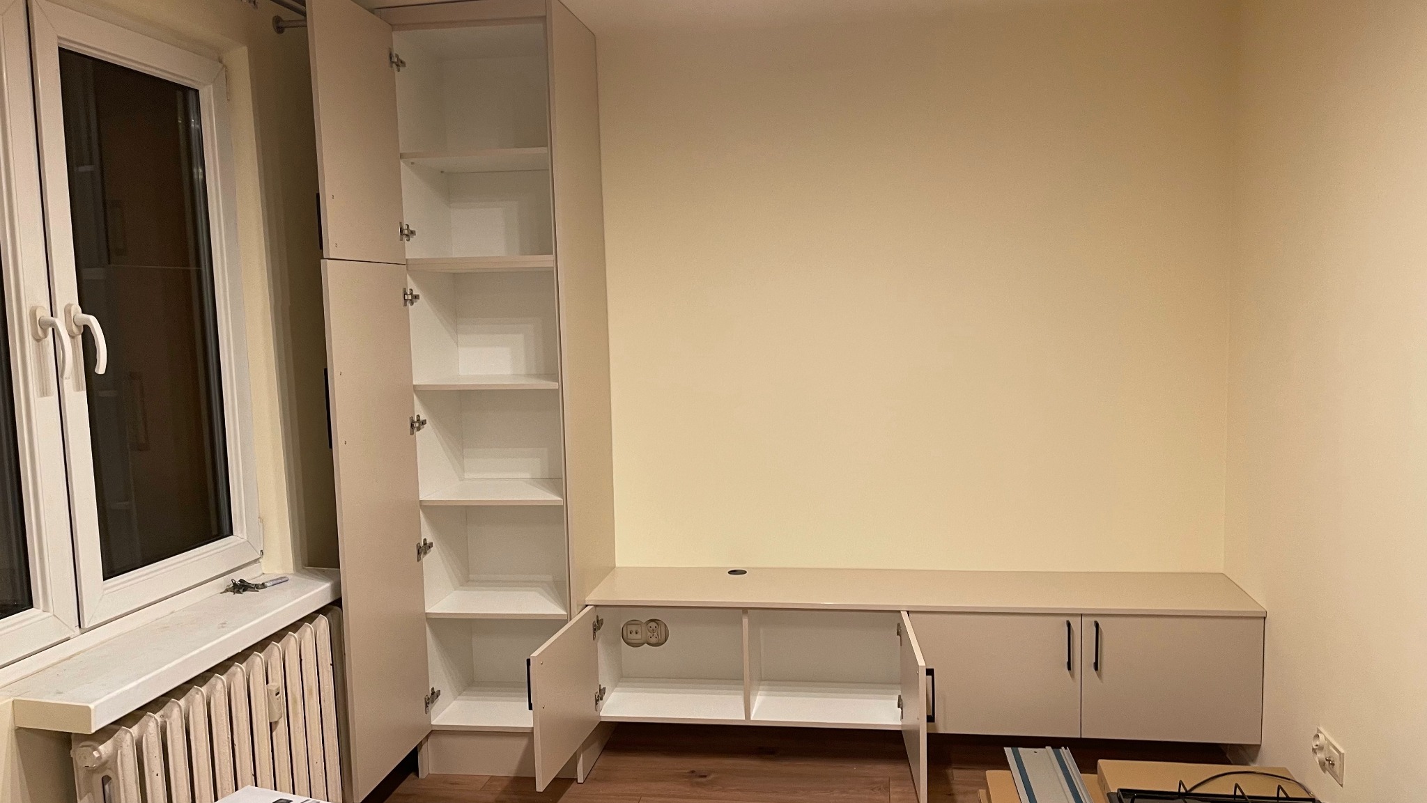 Biała zabudowa meblowa z szafkami i regałem w trakcie montażu, widoczne gniazdka elektryczne i narzędzia na podłodze.