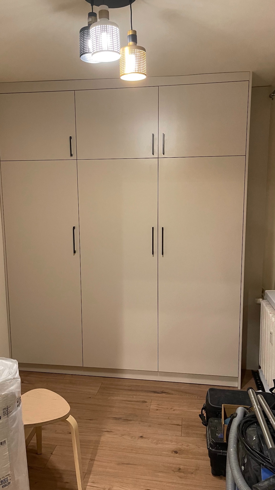 Jasna, trzydrzwiowa szafa na wymiar z minimalistycznymi uchwytami, oświetlona designerską lampą wiszącą z ażurowymi kloszami, widoczna w pokoju z drewnianą podłogą.