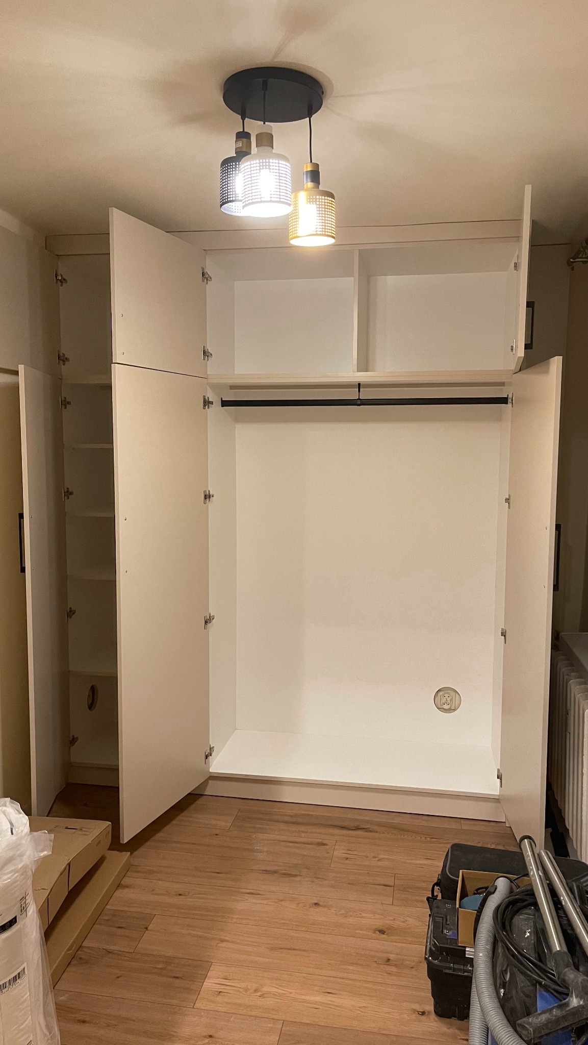 Wnętrze białej szafy wnękowej z otwartymi drzwiami, widoczny czarny drążek na ubrania, drewniana podłoga i nowoczesna lampa sufitowa