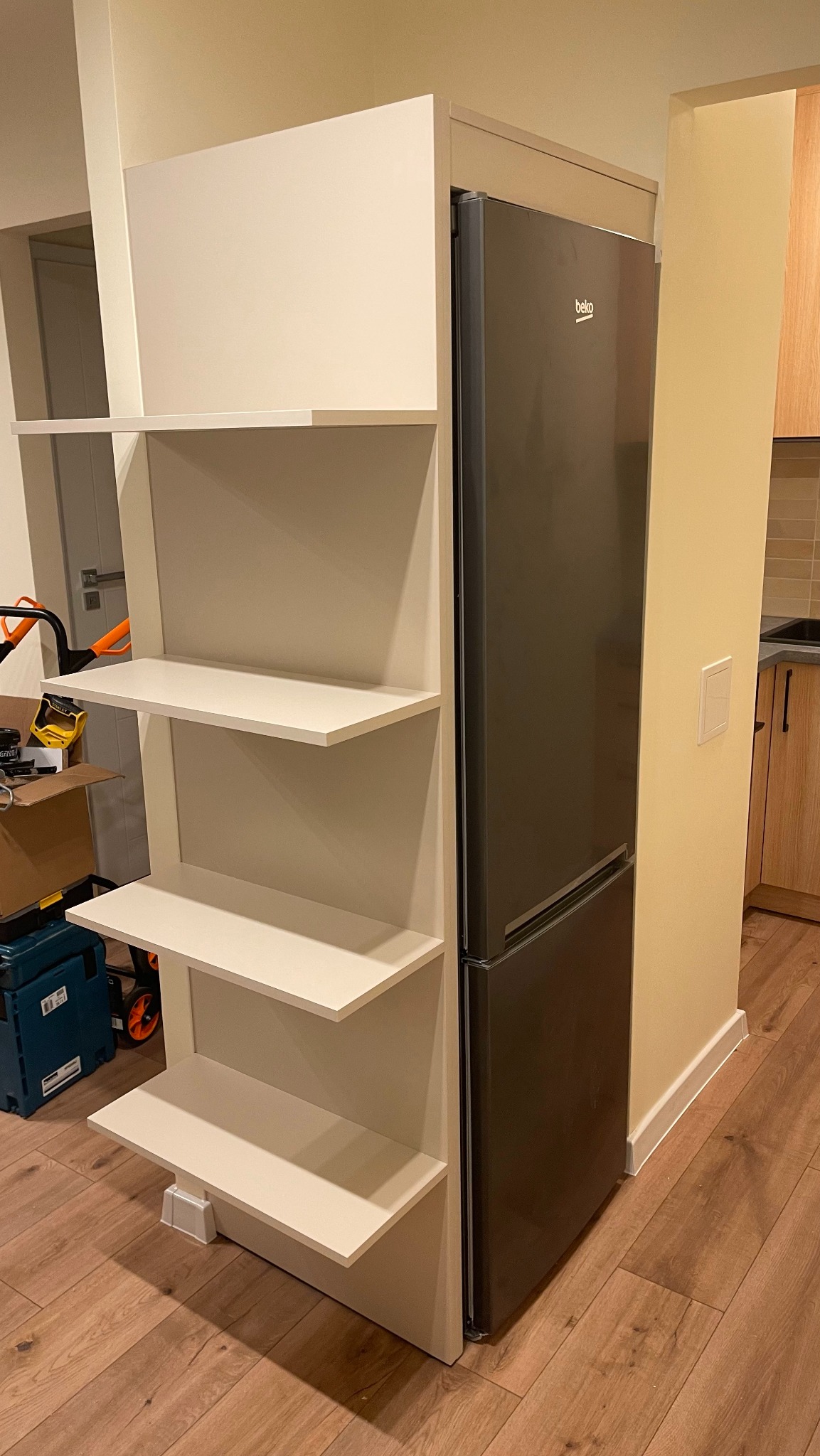 Biała szafka z półkami na wymiar, zabudowana obok lodówki Beko, widoczne narzędzia i kartony w tle.
