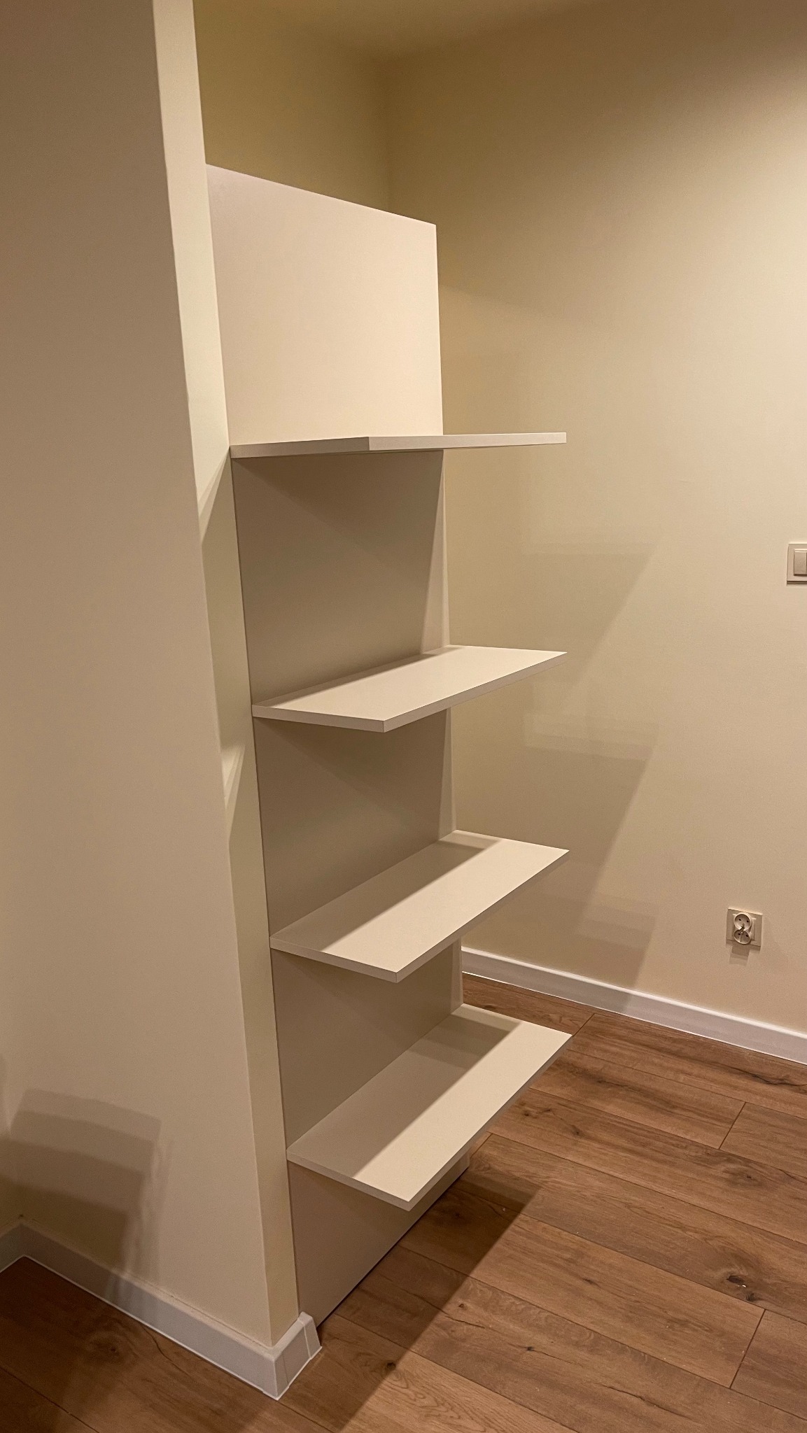Biała, minimalistyczna półka wnękowa z czterema poziomami, wbudowana w ścianę, na tle drewnianej podłogi i kremowych ścian.