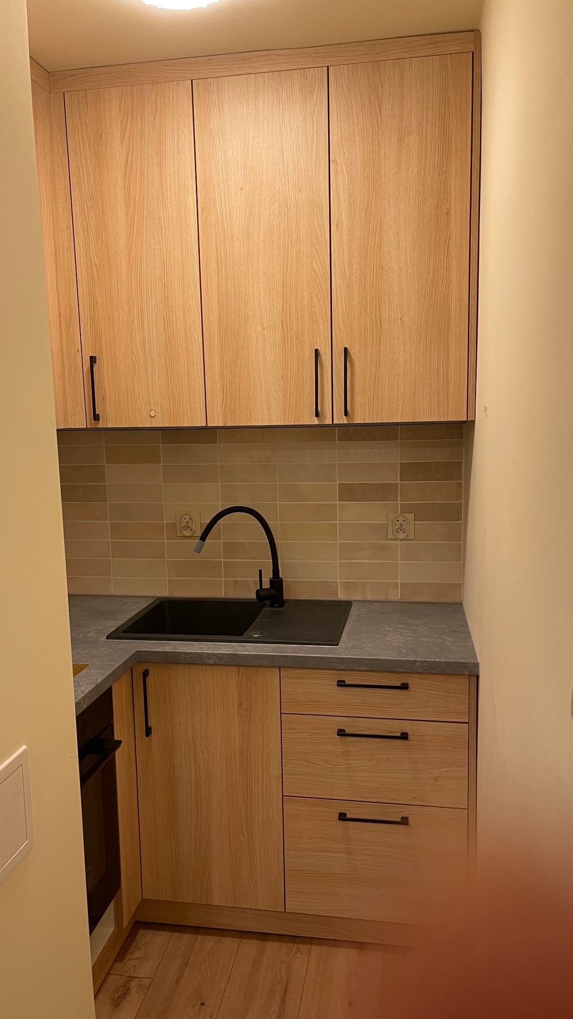 Jasna, drewniana zabudowa kuchenna z czarnym zlewem i baterią; płytki w odcieniach beżu na ścianie, minimalistyczne czarne uchwyty.