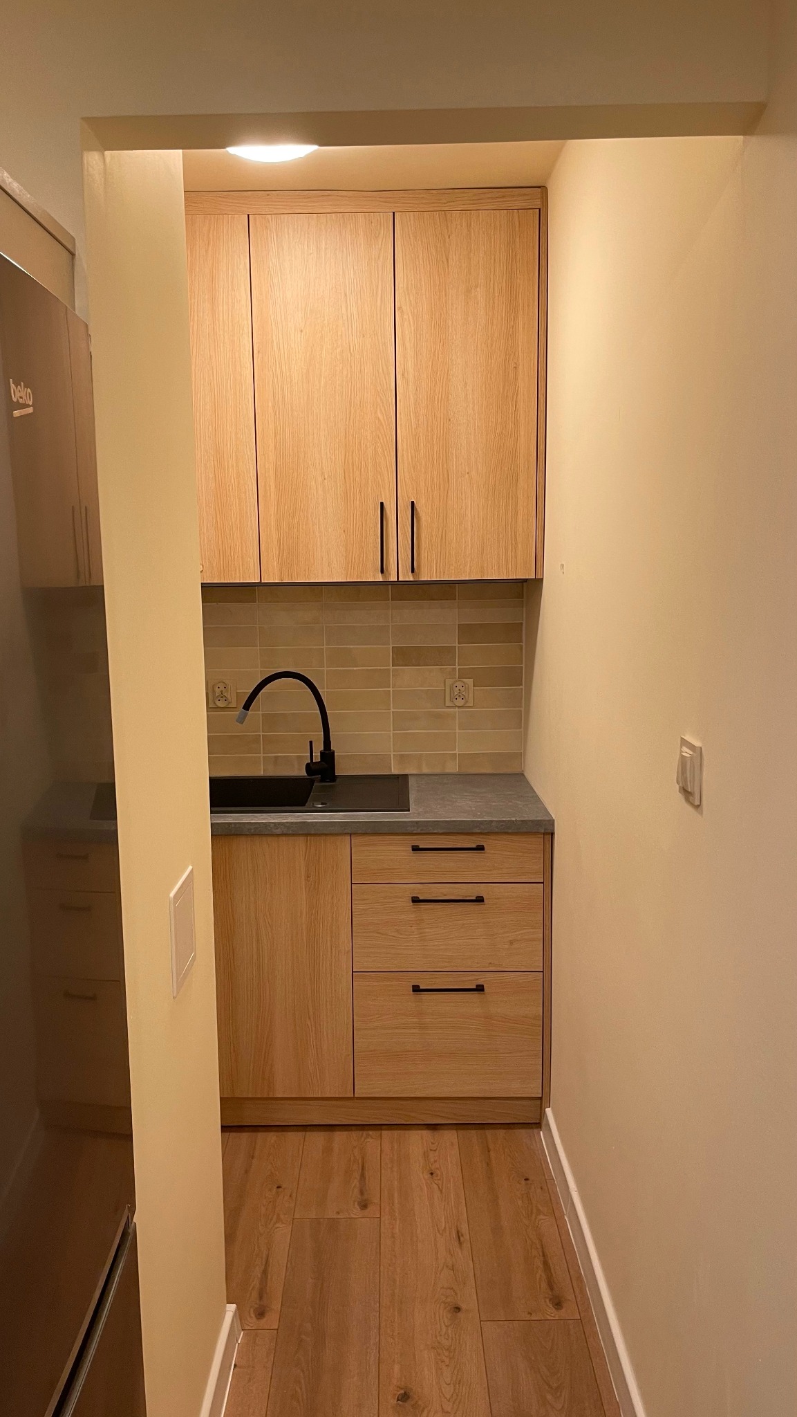 Wąska kuchnia z drewnianymi frontami, czarną armaturą i szarym blatem, widok z perspektywy wejścia do pomieszczenia.