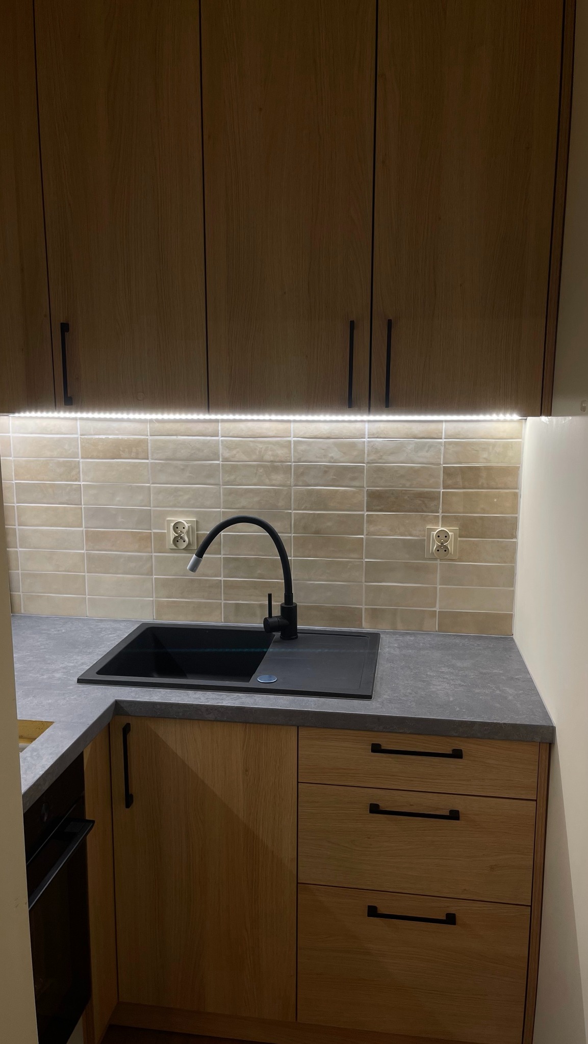 Fragment aranżacji kuchni z drewnianymi szafkami, czarnym zlewem i baterią, oświetleniem LED podszafkowym i beżowymi płytkami na ścianie.