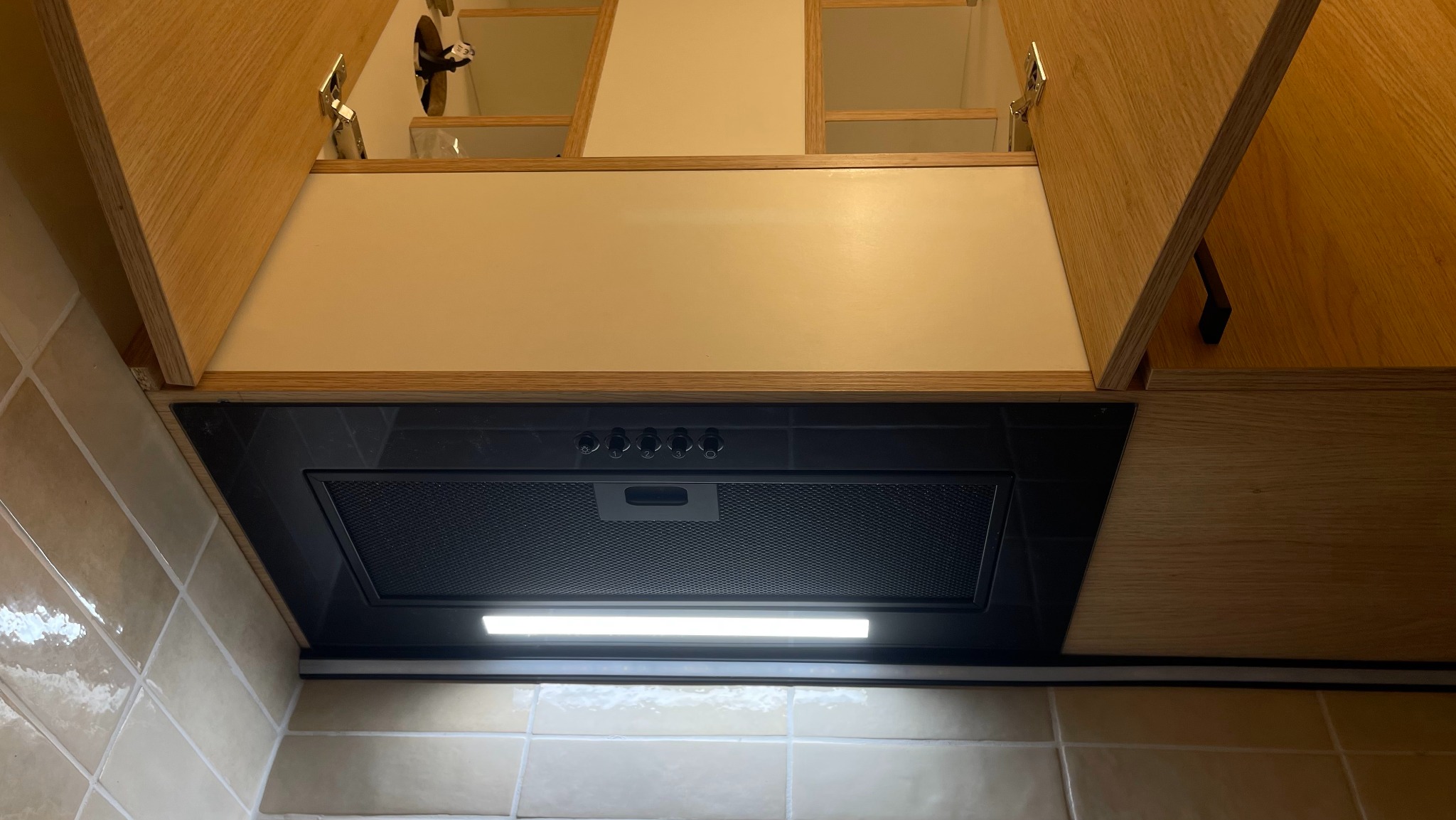 Zamontowany okap podszafkowy w zabudowie kuchennej z otwartymi szafkami z jasnego drewna i beżowymi płytkami na ścianie.