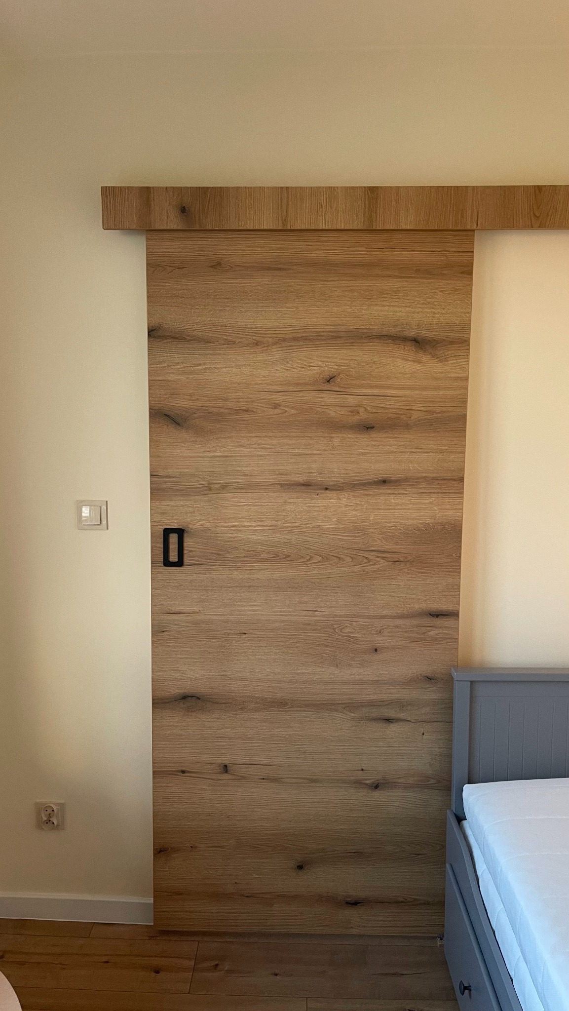 Drewniane drzwi przesuwne z czarnym uchwytem, zamontowane na ścianie w sypialni, widoczny fragment łóżka.