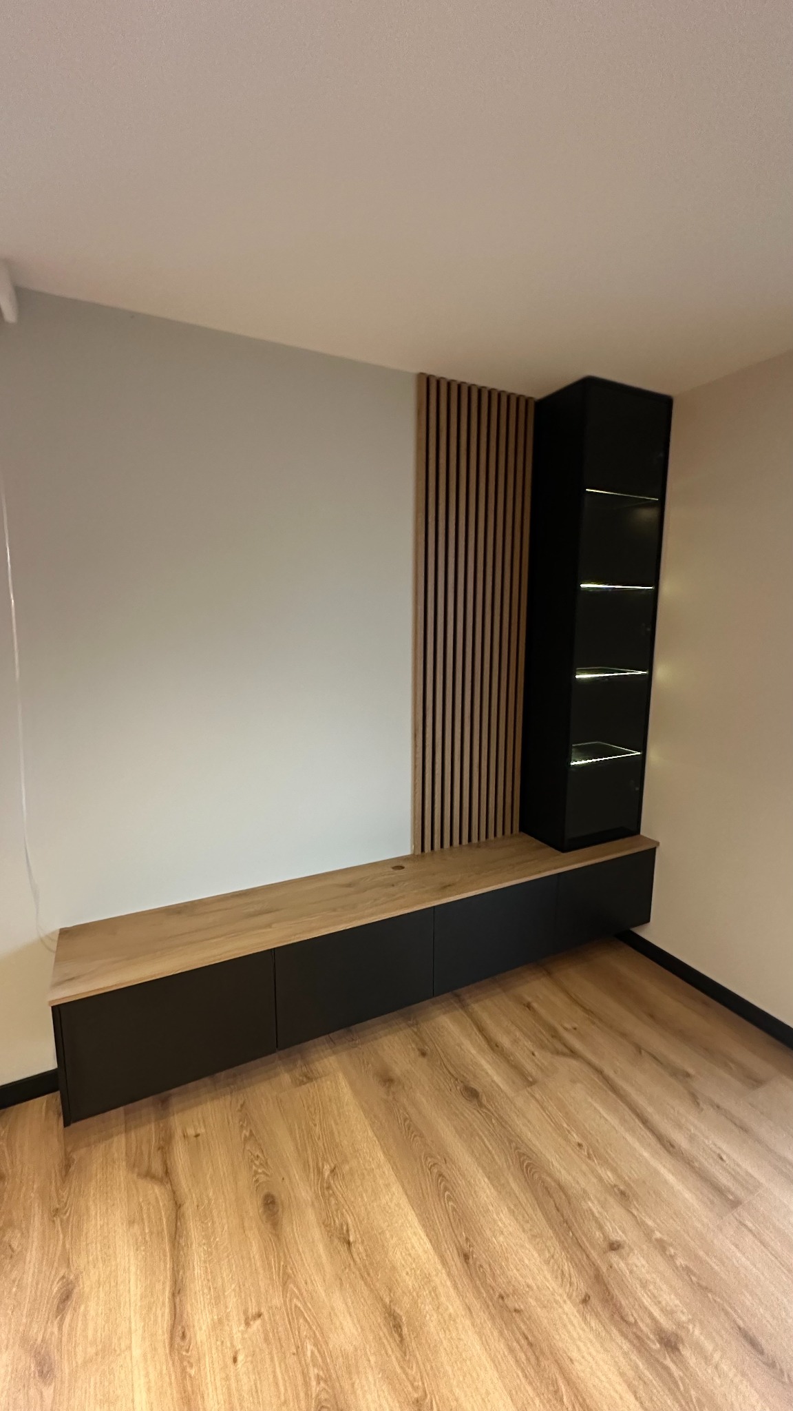 Nowoczesna zabudowa ściany z czarnymi szafkami i drewnianym blatem, uzupełniona pionowymi panelami dekoracyjnymi i podświetlaną witryną.