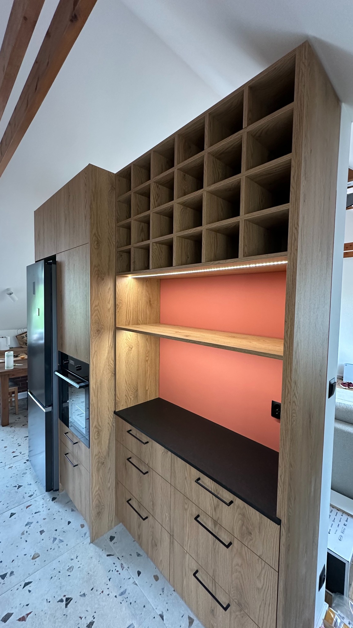 Drewniana zabudowa kuchenna z szafkami, szufladami z czarnymi uchwytami i otwartą półką z oświetleniem LED na tle koralowej ściany oraz regałem na wino. Obok stoi lodówka w zabudowie. Na podłodze...