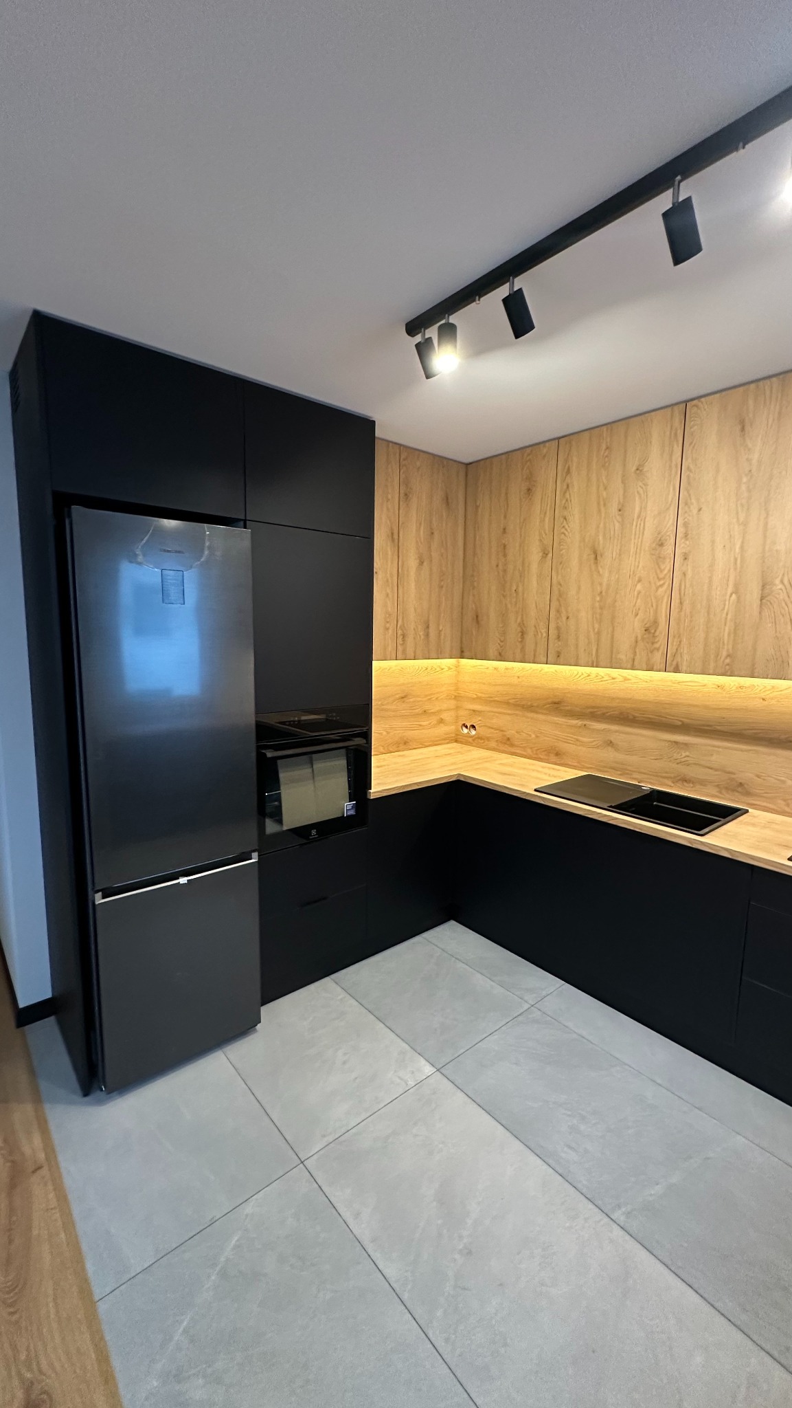 Nowoczesna kuchnia z czarnymi frontami i drewnopodobnymi szafkami górnymi, oświetlenie LED pod szafkami, szara podłoga z płytek.