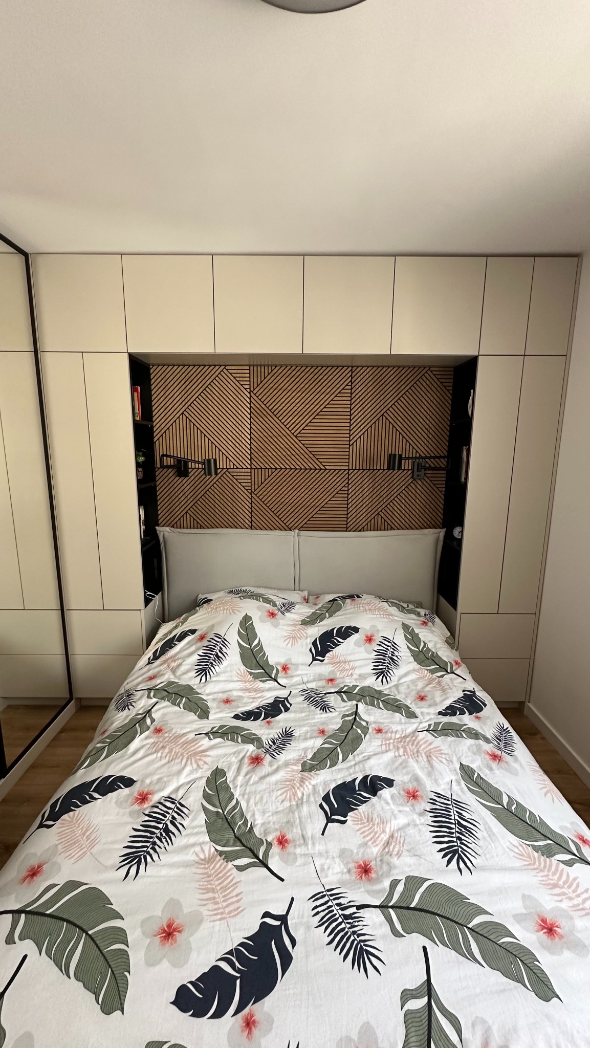 Sypialnia z łóżkiem, wezgłowiem z drewnianych paneli dekoracyjnych i szafą wnękową po bokach, oświetlenie punktowe, pościel w liście.