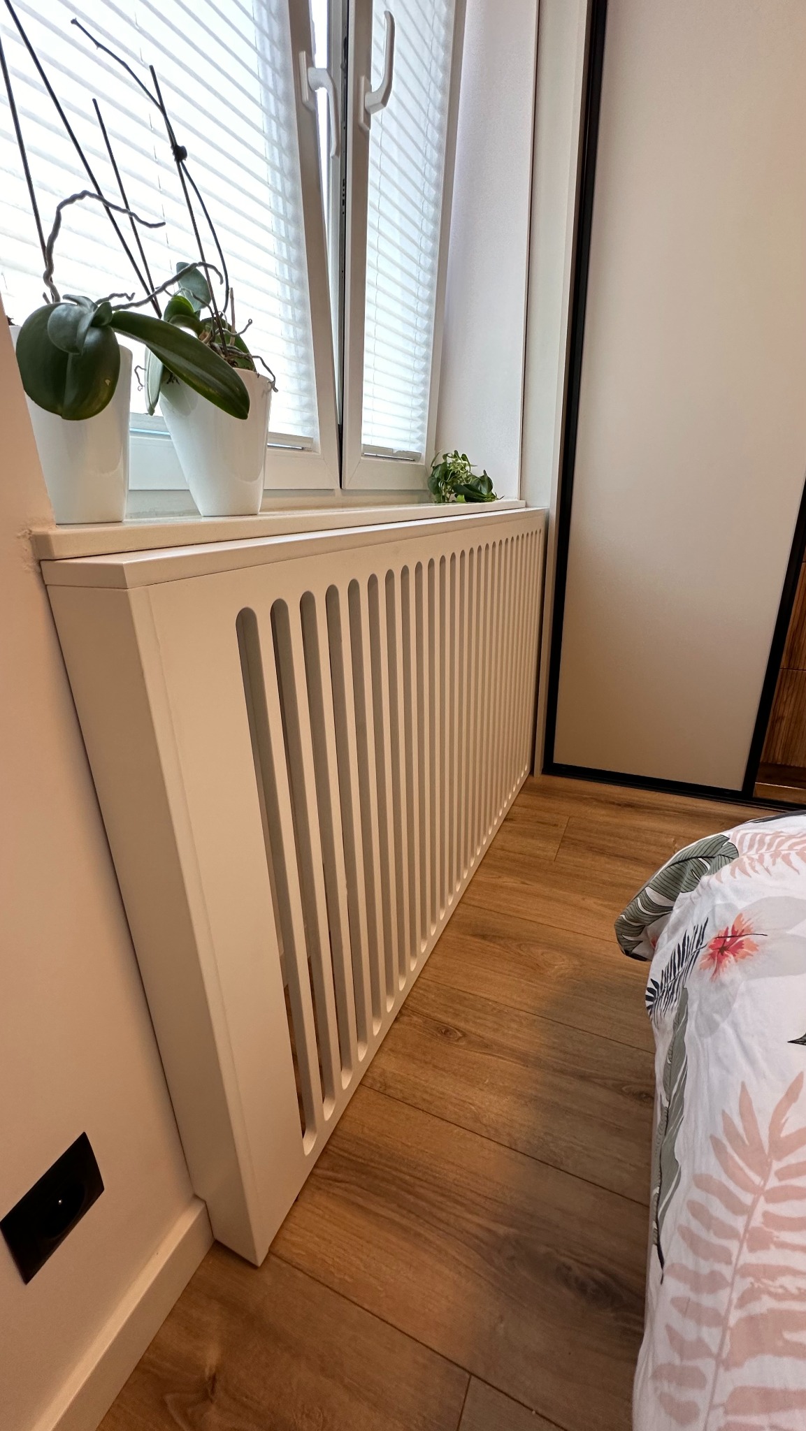 Biała obudowa grzejnika z pionowymi szczelinami, dopasowana do koloru parapetu i ramy okiennej, z roślinami w doniczkach na parapecie, drewnopodobna podłoga.