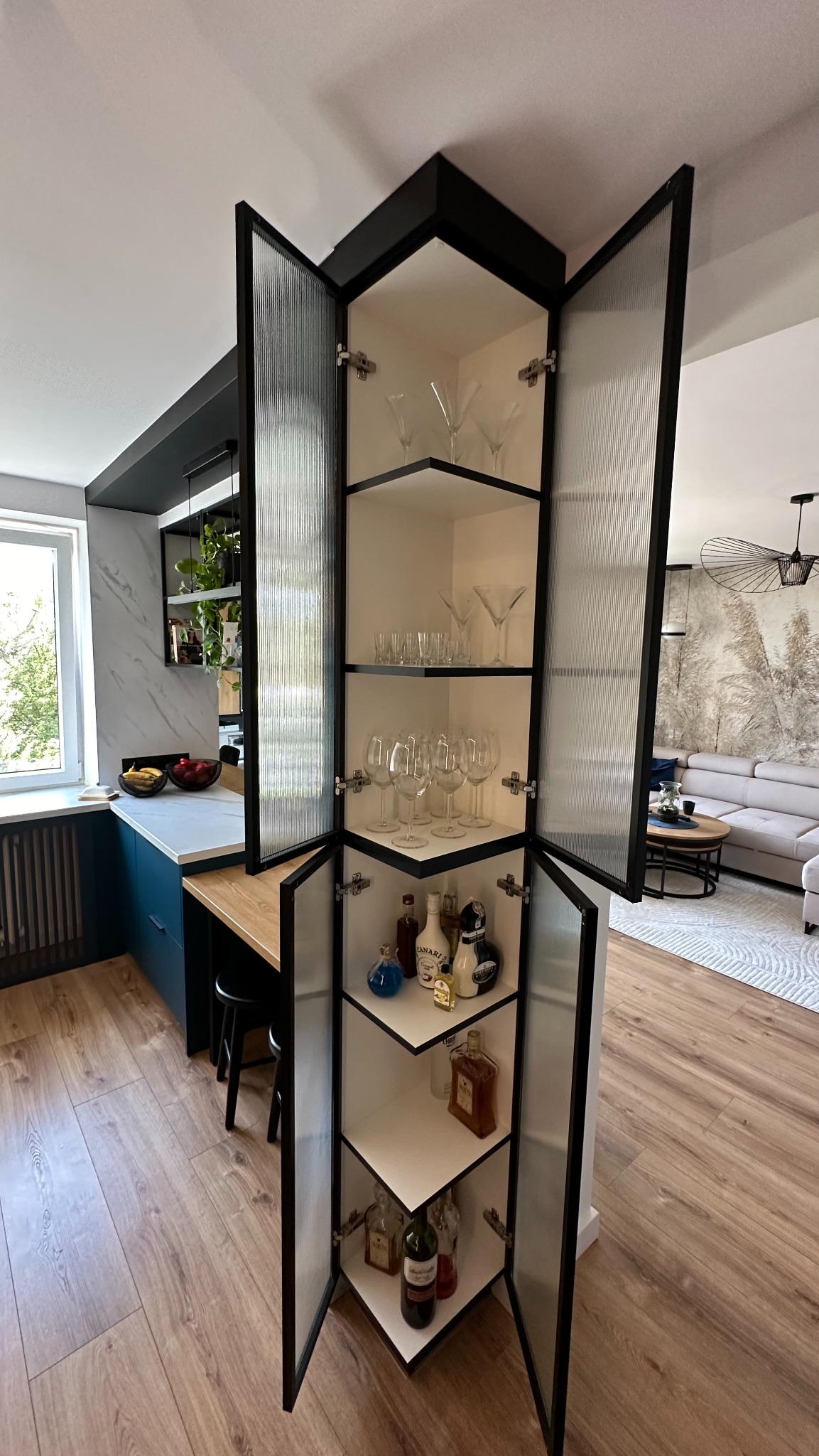 Wysoka narożna szafka z ryflowanymi szklanymi drzwiczkami, wypełniona szkłem barowym i butelkami alkoholu, widoczna w nowoczesnym wnętrzu kuchni z salonem. Czarny obrys szafki kontrastuje z białymi...