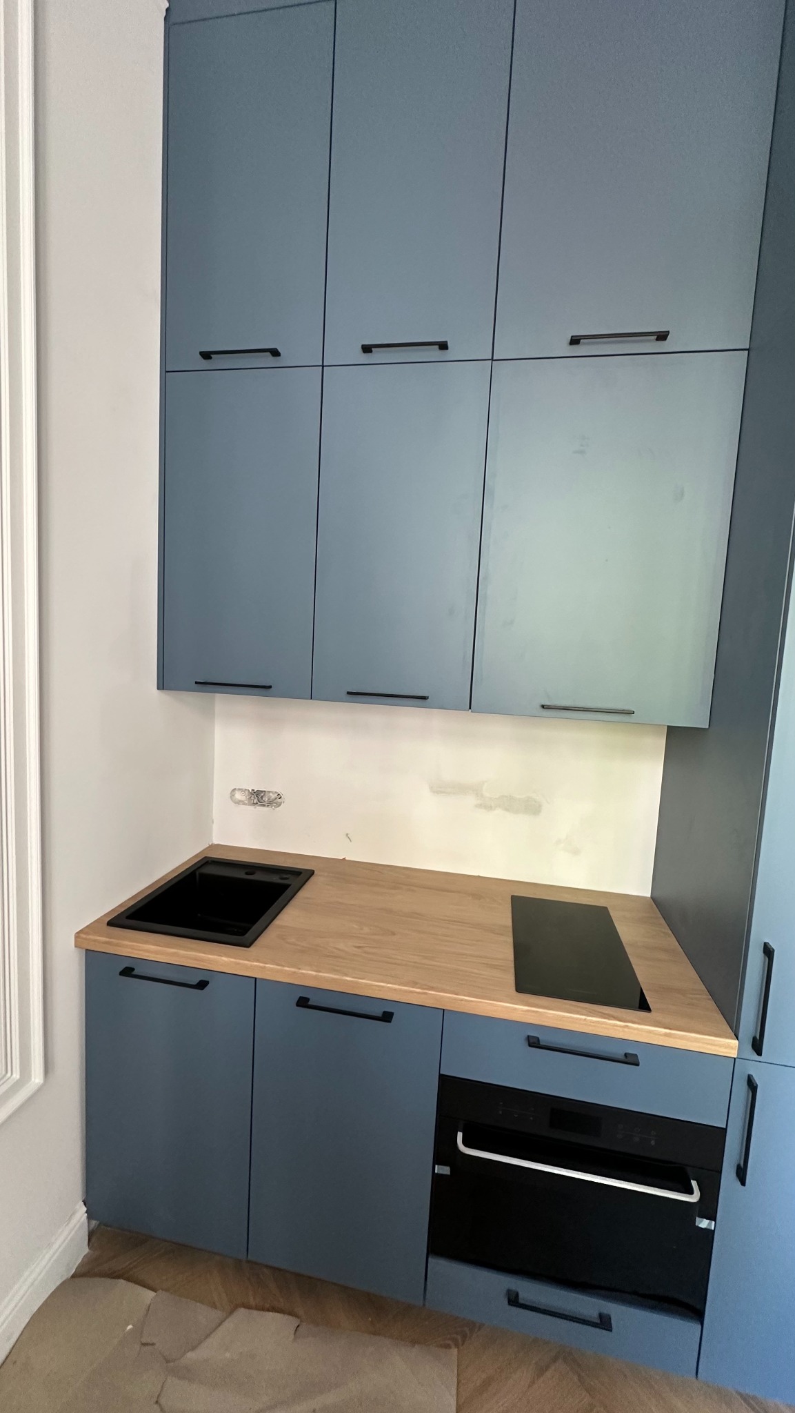 Niebieska zabudowa kuchenna z drewnianym blatem, czarnym zlewem i płytą indukcyjną, widoczne ślady po instalacji na ścianie, minimalistyczne uchwyty.