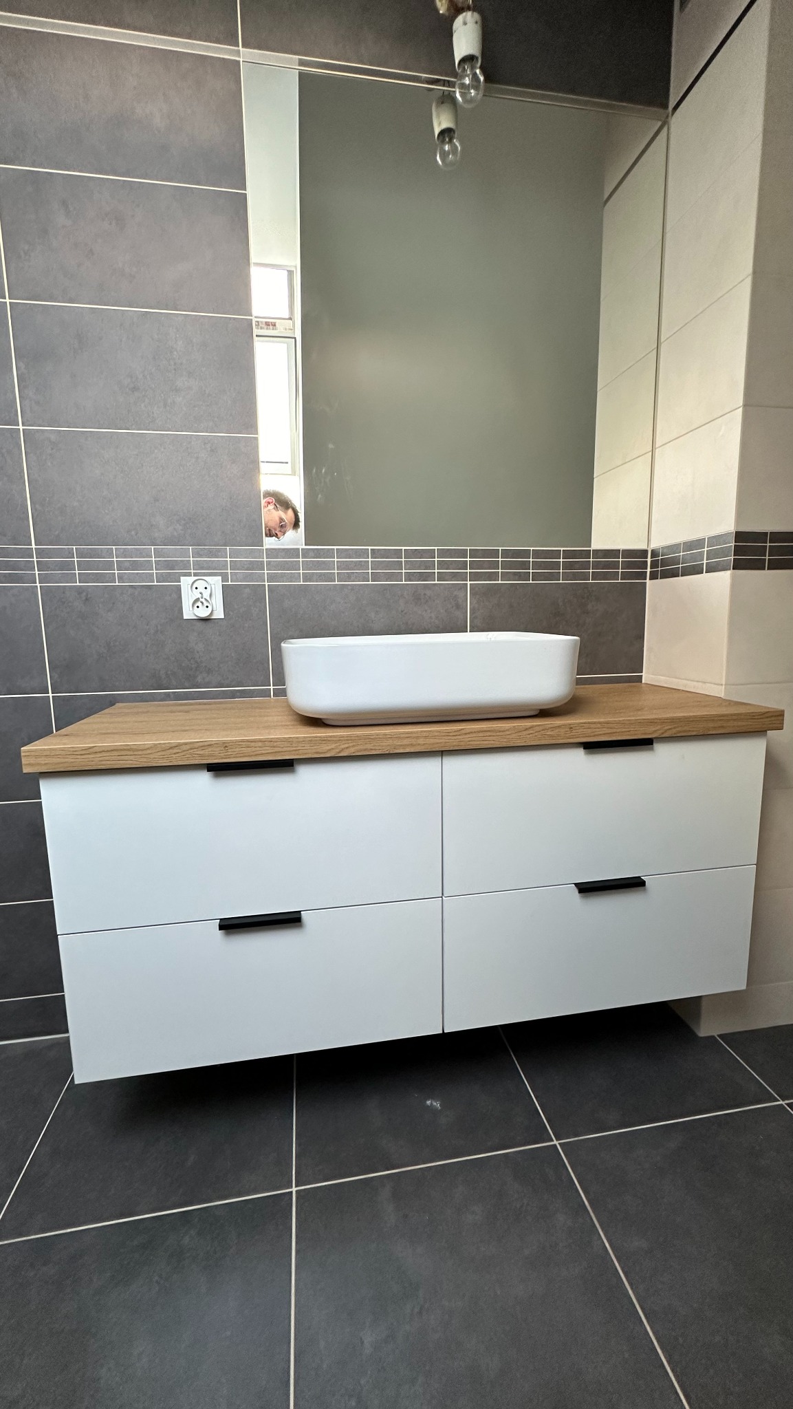 Nowoczesna łazienka z umywalką nablatową na szafce z białymi frontami i drewnianym blatem, ciemne płytki podłogowe i ścienne, widoczne odbicie osoby w lustrze.