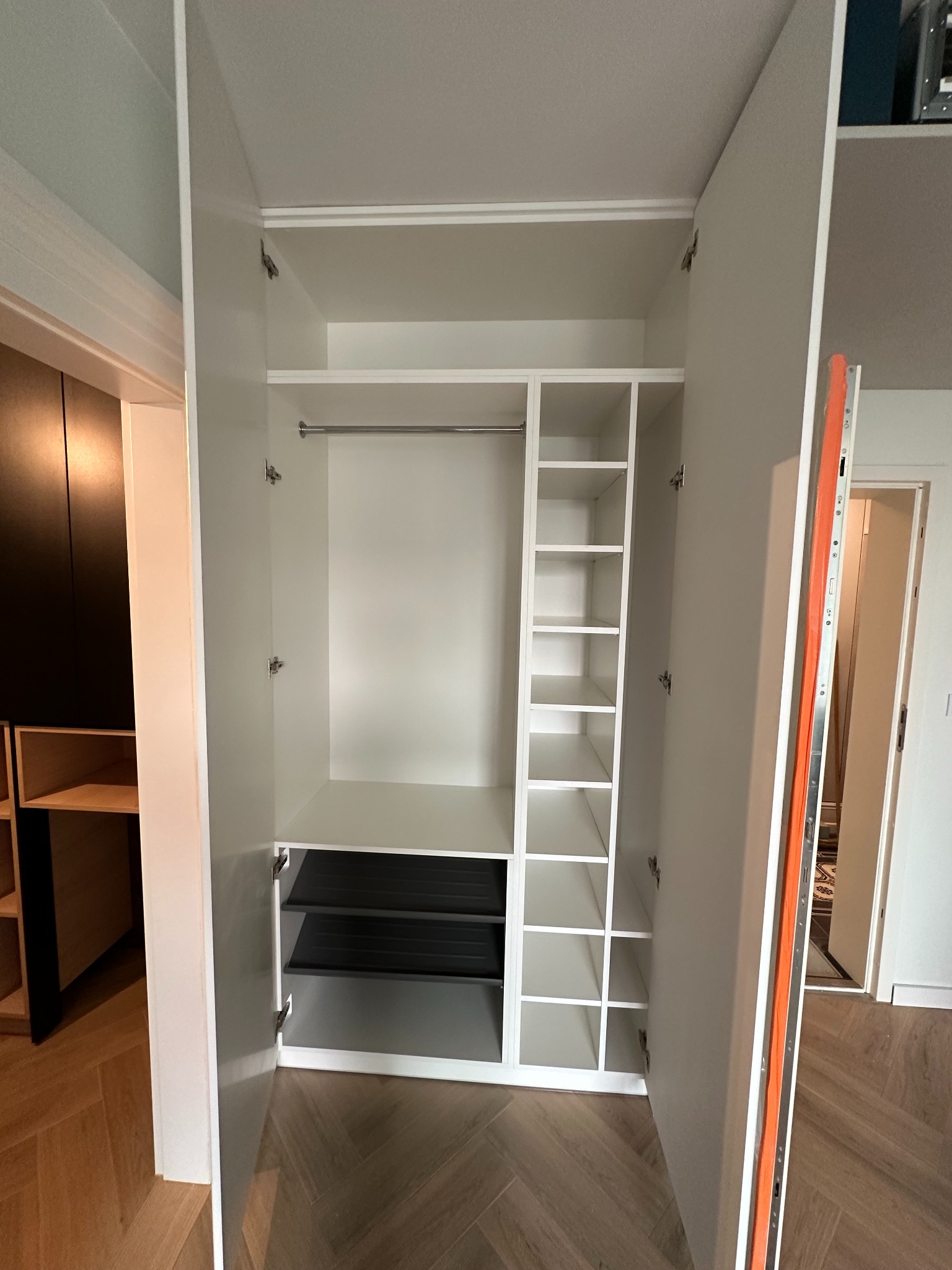 Wnętrze białej szafy wnękowej z drzwiami na zawiasach, widoczne półki, drążek i wysuwane kosze w kolorze czarnym.