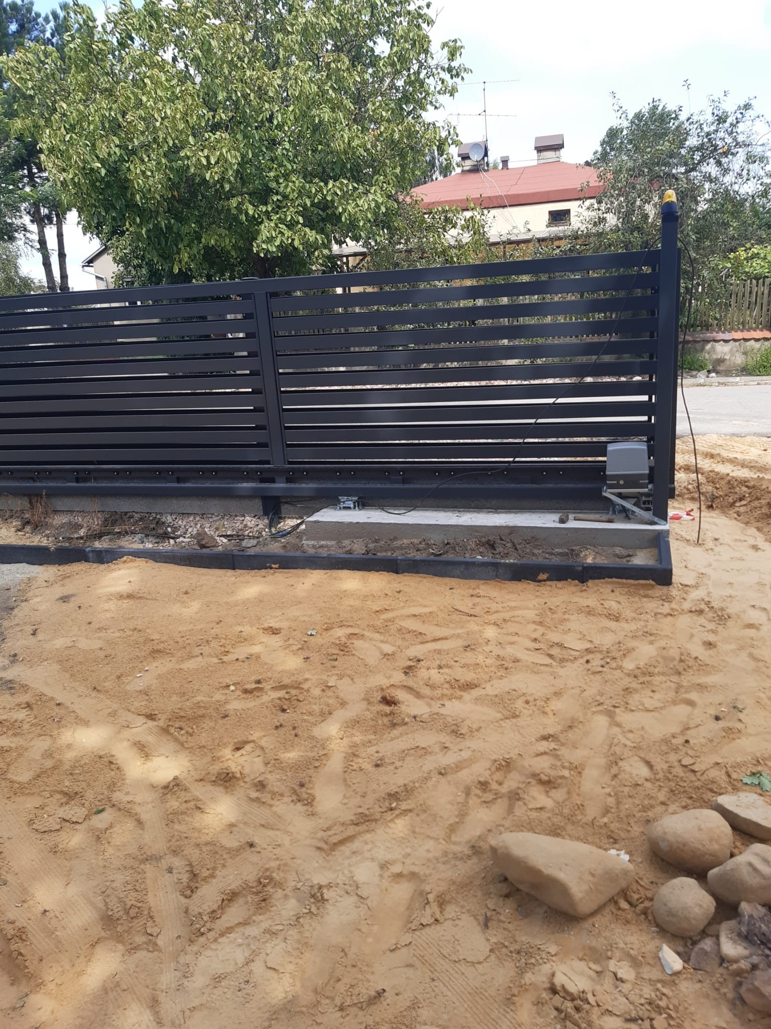 Nowoczesna, przesuwna brama wjazdowa z poziomych profili w kolorze antracytowym, zamontowana na betonowej podstawie z obrzeżami, widoczny automat do bramy, teren wokół w trakcie prac ziemnych.