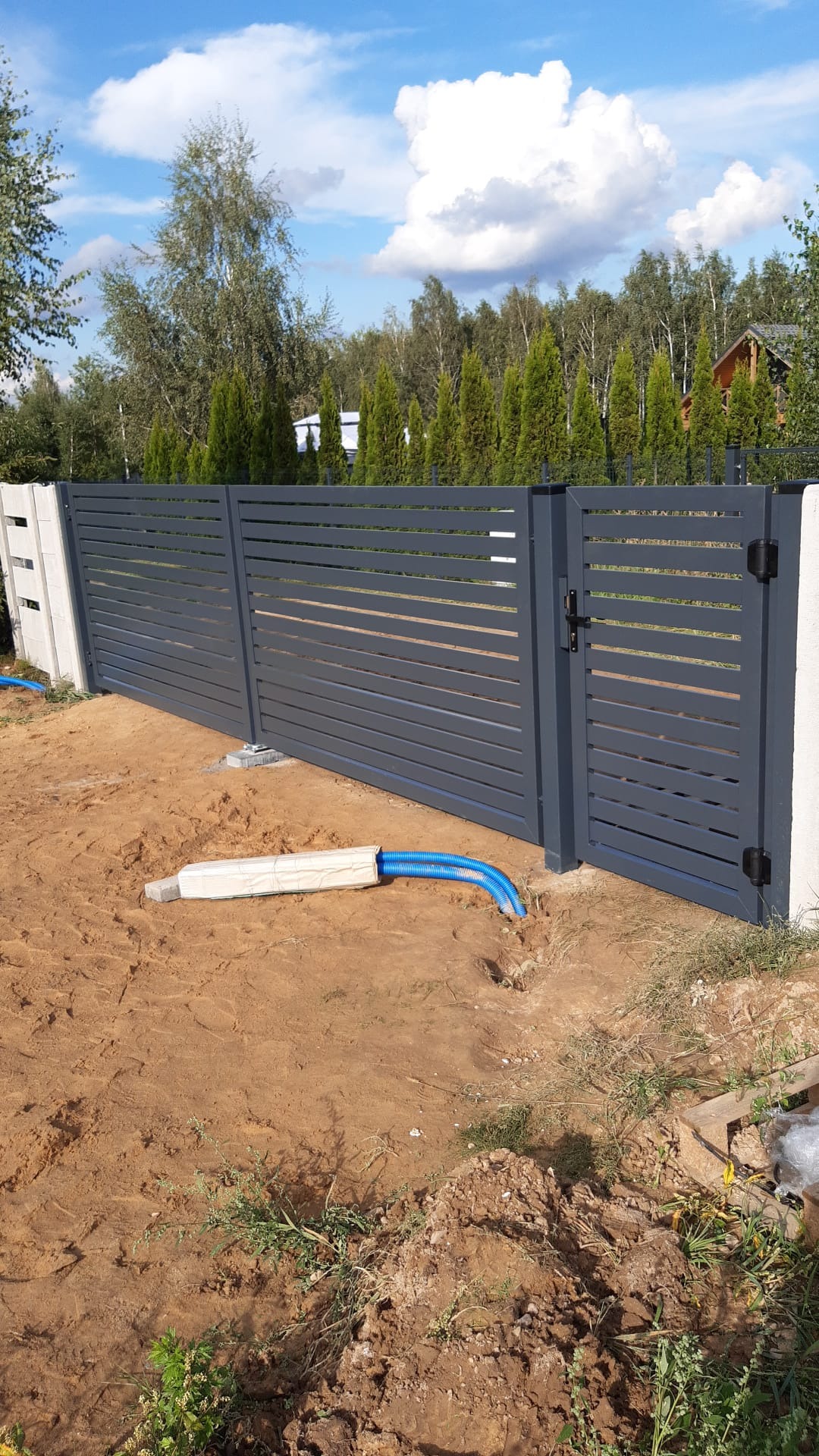 Nowoczesna, pozioma, szara brama wjazdowa i przyległe ogrodzenie panelowe z betonowymi słupkami, widoczne niebieskie rury instalacyjne w ziemi i częściowo zasypane piaskiem, w tle drzewa i błękitne...