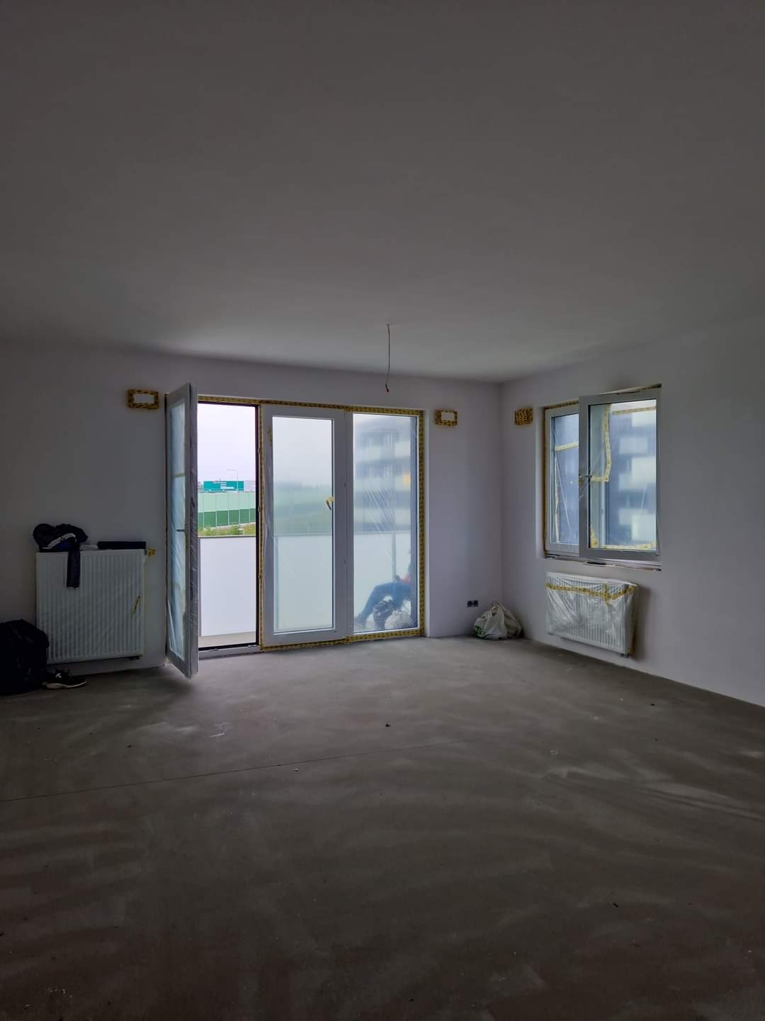 Wnętrze w stanie deweloperskim z przygotowanym podłożem, zabezpieczonymi oknami i grzejnikami, widoczne ślady po szpachlowaniu na ścianach, otwarte drzwi balkonowe.