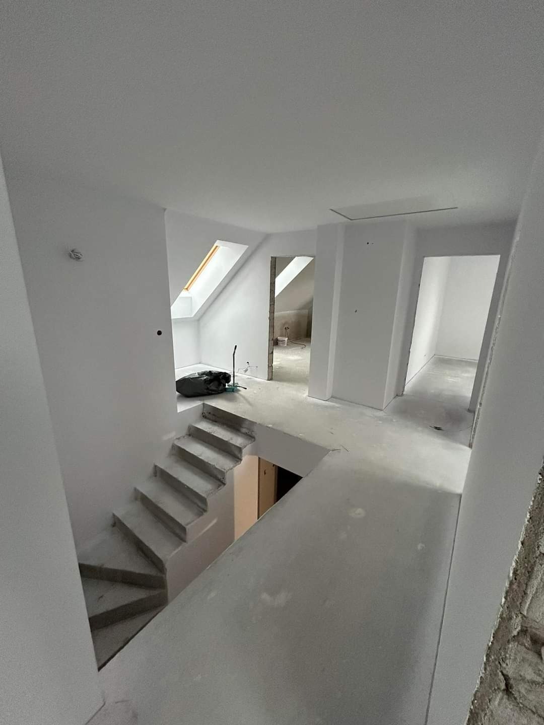 Wnętrze w trakcie remontu: białe ściany, surowe schody betonowe, widoczne niedoskonałości i przygotowania do dalszych prac wykończeniowych, okno dachowe w oddali.