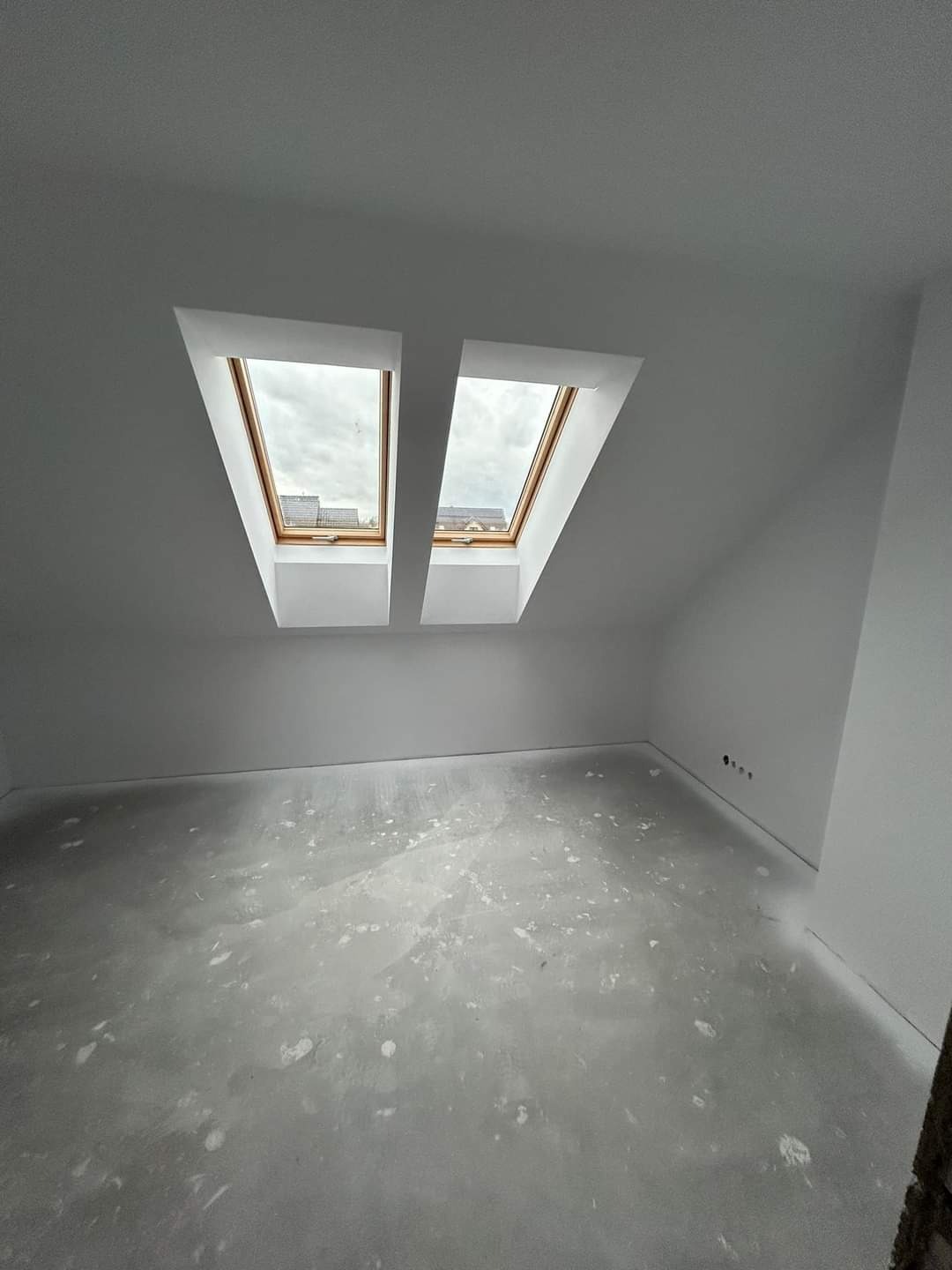 Poddasze z dwoma oknami dachowymi, ściany pomalowane na biało, podłoga w trakcie przygotowania, widoczne ślady po szpachlowaniu.