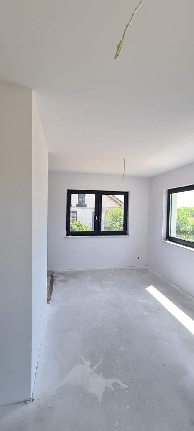Pomalowane na biało wnętrze pokoju z dwoma oknami w czarnych ramach, betonowa podłoga z plamami farby, widoczne kable zwisające z sufitu.