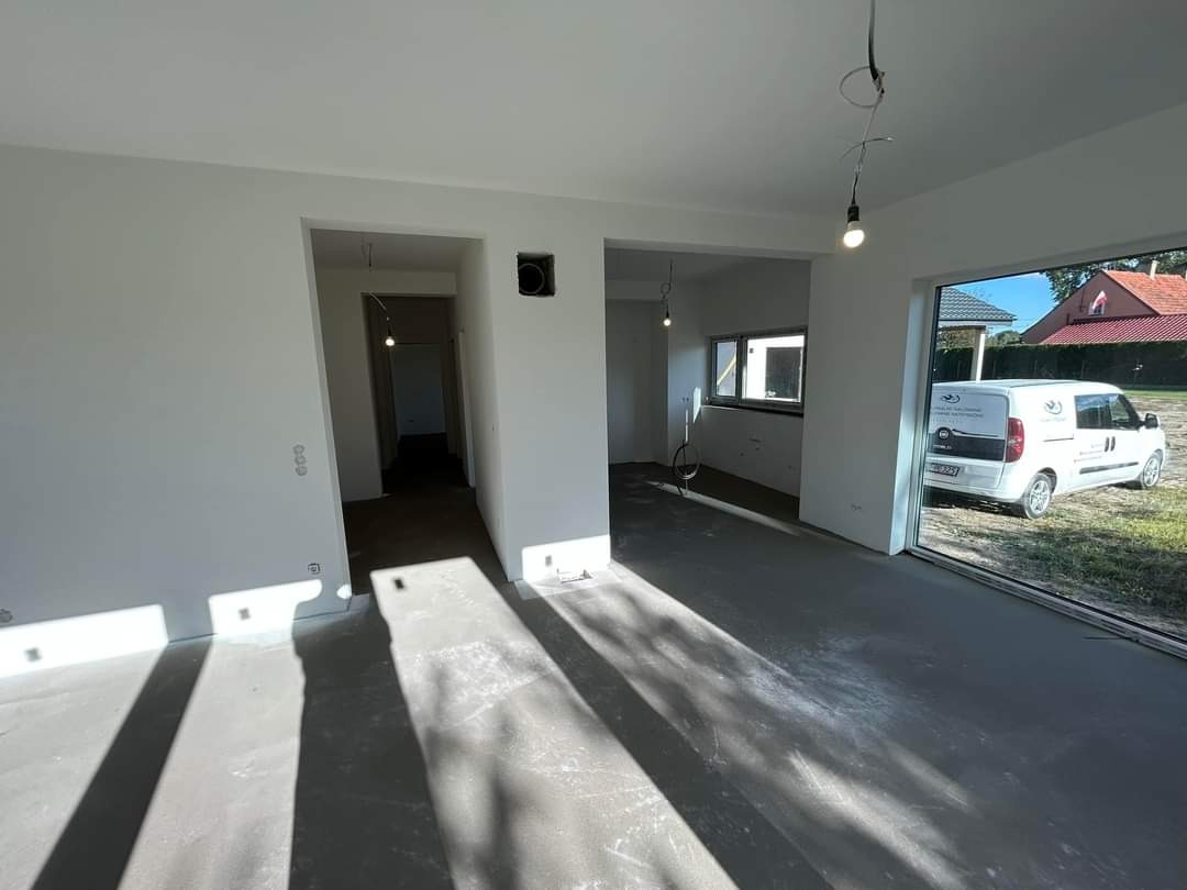 Wnętrze świeżo wykończonego pomieszczenia z gładkimi, białymi ścianami i szarą podłogą, widoczne przygotowane punkty oświetleniowe, na zewnątrz przez okno widoczny samochód dostawczy firmy.