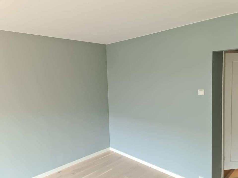 Świeżo pomalowany pokój w odcieniach szarości i bieli, widoczny fragment podłogi z jasnych paneli oraz włącznik światła na ścianie.