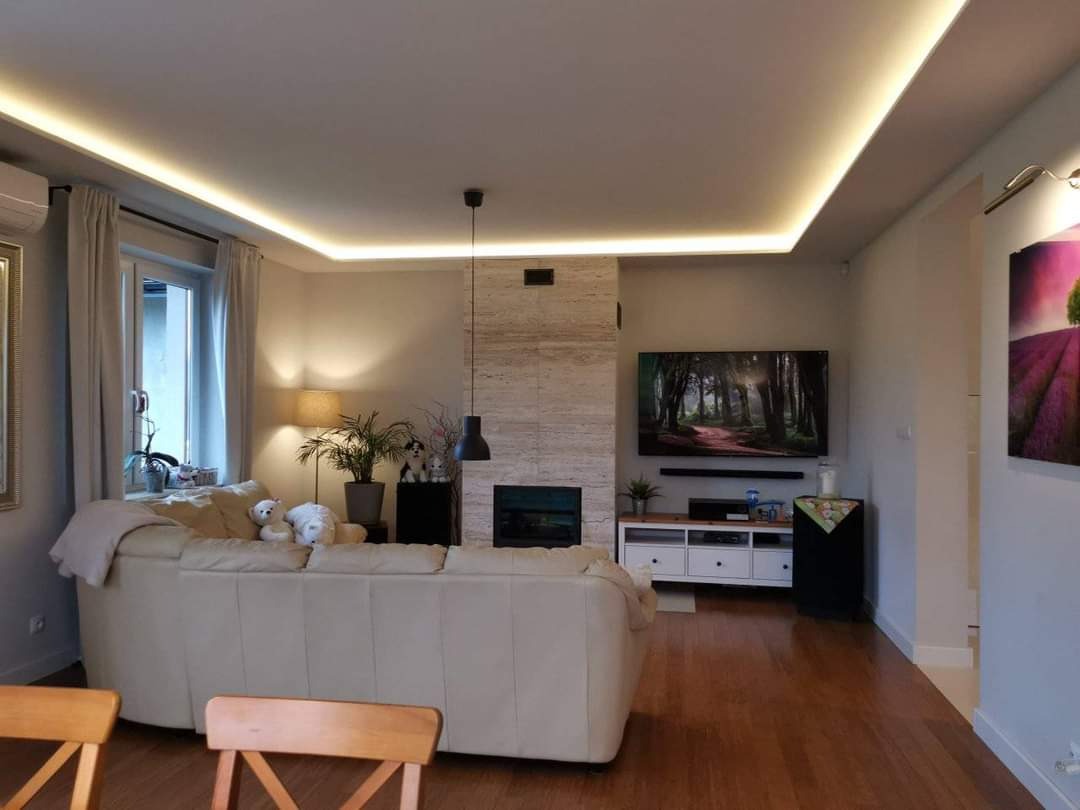 Salon z oświetleniem LED podsufitowym, parkietem na podłodze, kominkiem obłożonym kamieniem dekoracyjnym i telewizorem na ścianie.