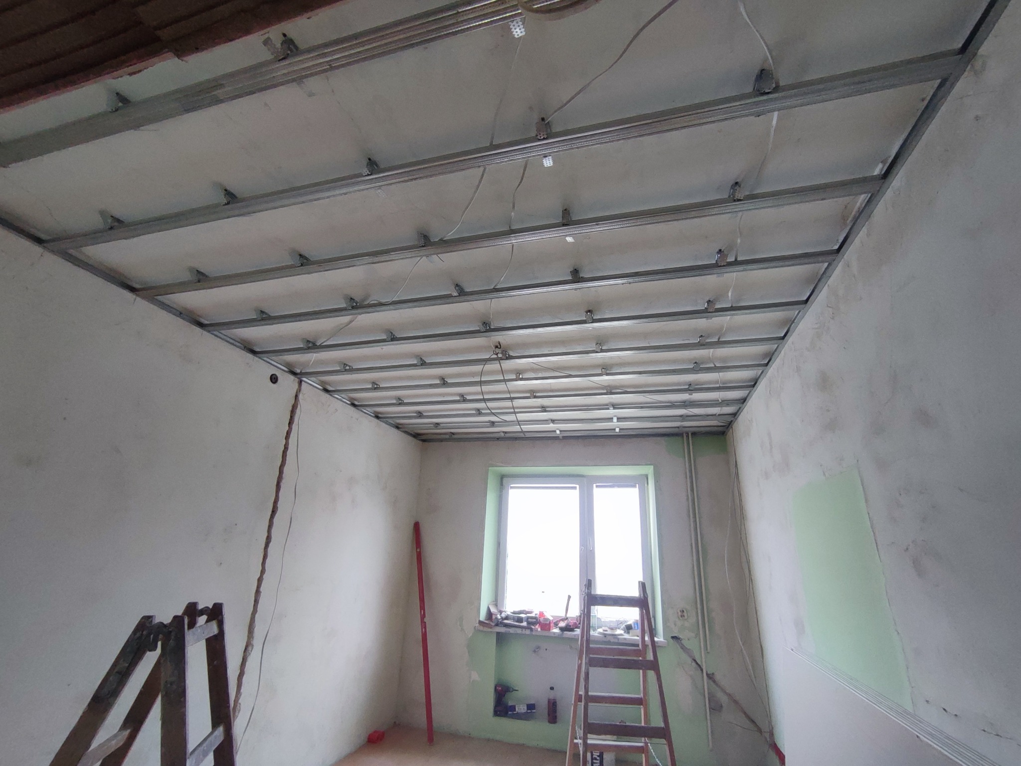 Stelaż sufitu podwieszanego z metalowych profili w surowym wnętrzu, widoczne przewody, drabina i narzędzia przy oknie.