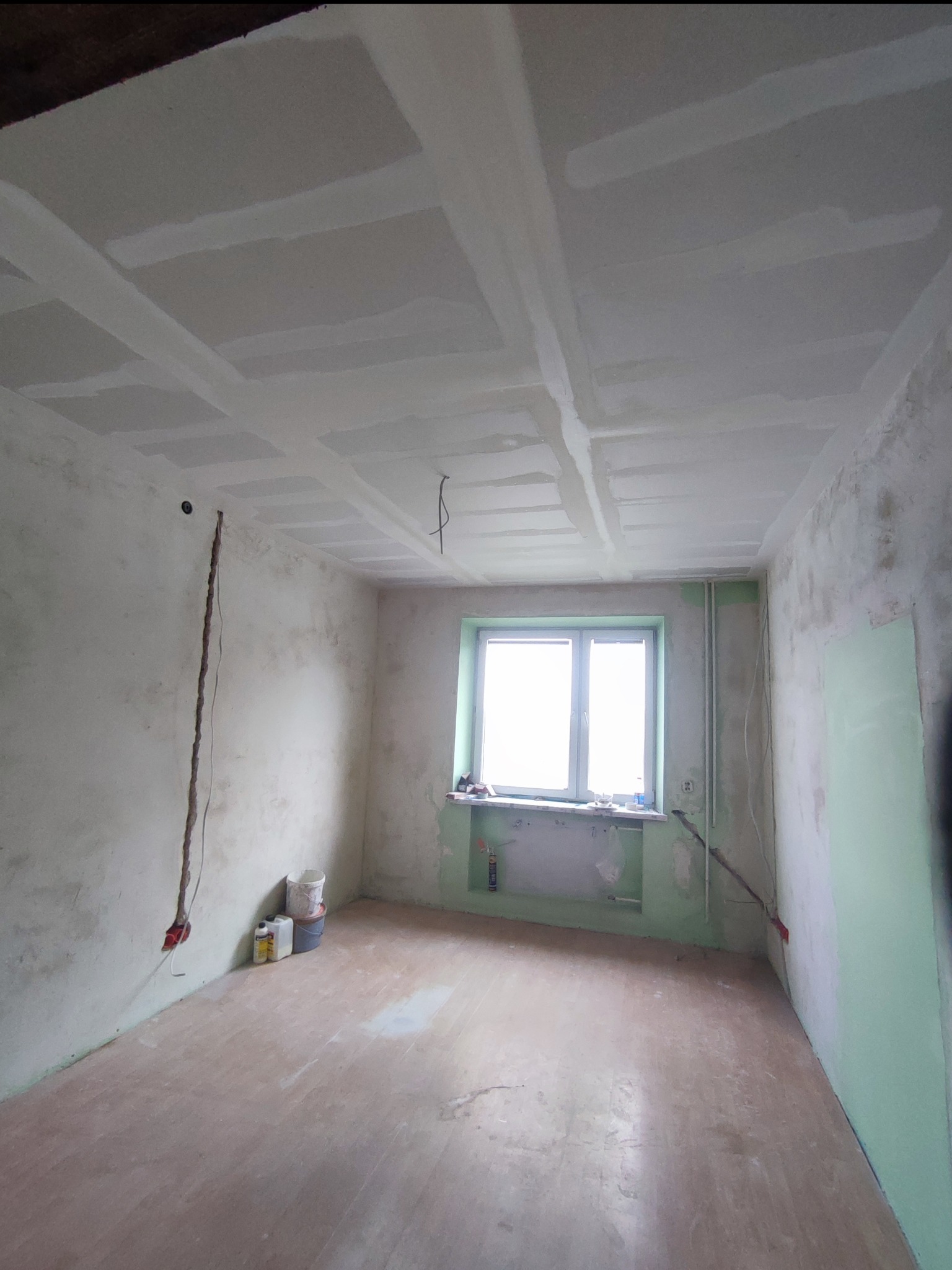 Pokój w trakcie remontu: surowy sufit z płyt kartonowo-gipsowych z widocznymi łączeniami, ściany w stanie surowym, widoczne puszki elektryczne i przewody, okno z parapetem, podłoga przygotowana...