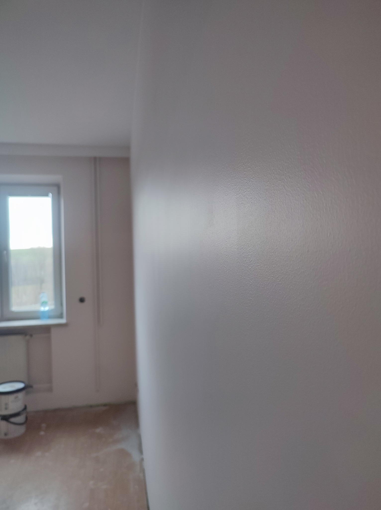 Świeżo pomalowana ściana w pokoju, widoczny fragment okna i wiadro farby na podłodze, delikatna faktura farby odbija światło.