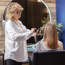 Fryzjerka w białej bluzce i okularach prostuje włosy klientce siedzącej w czarnym fotelu, widok z tyłu, okrągłe lustro z podświetleniem w tle na drewnianej ścianie.