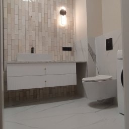 RemontujemyDlaCiebie - Nowoczesna łazienka z beżowymi, pionowo ułożonymi płytkami na ścianie, wiszącą szafką z umywalką, podwieszanym WC i marmurową podłogą.