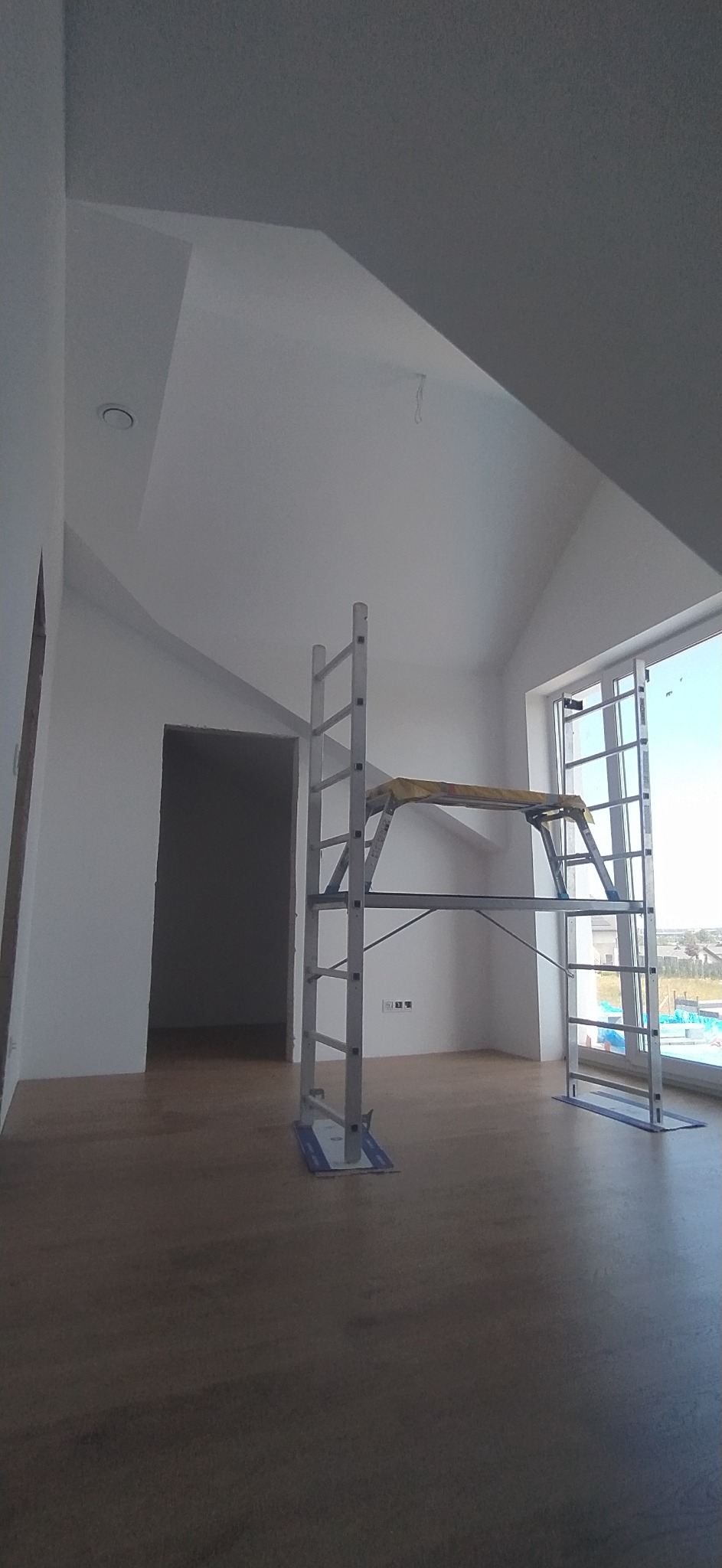 Poddasze po remoncie, świeżo pomalowane na biało, widoczne rusztowanie malarskie, drewniana podłoga, okno z widokiem na zabudowę jednorodzinną.