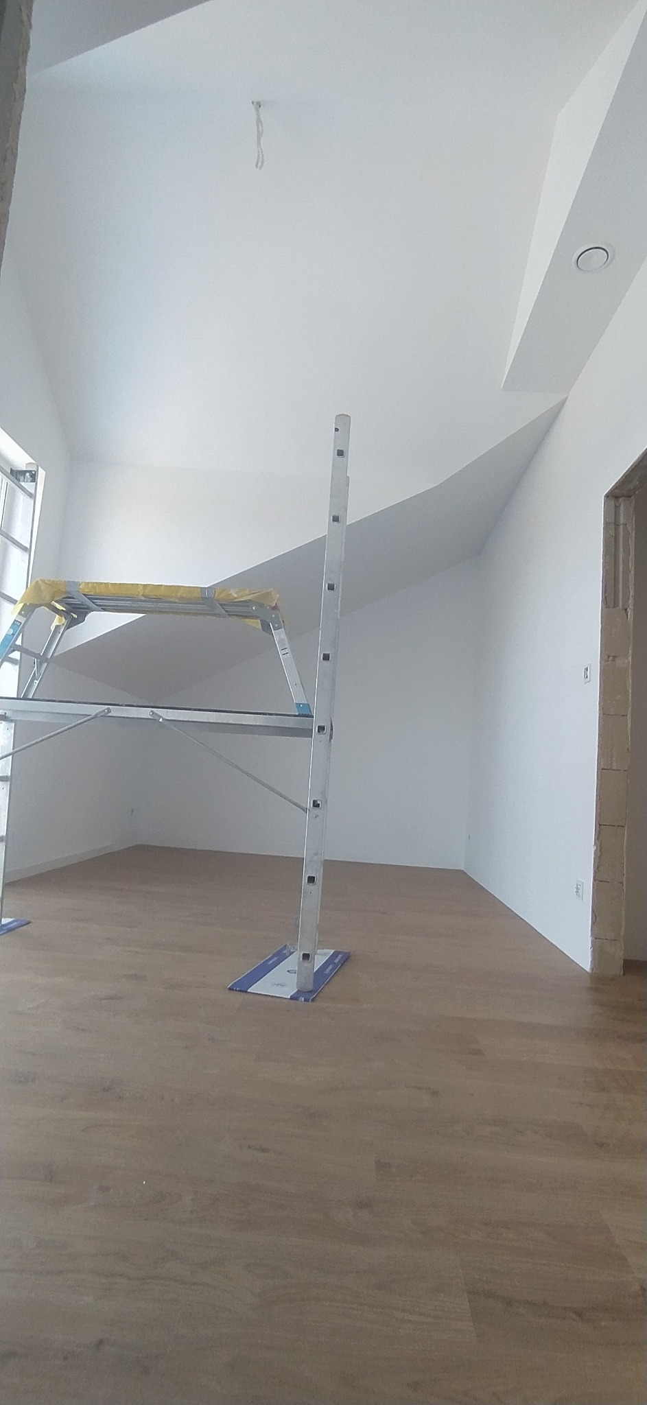 Wnętrze pokoju z panelami podłogowymi, świeżo pomalowanymi białymi ścianami i sufitem, z widocznym rusztowaniem malarskim z osłoniętą platformą roboczą.