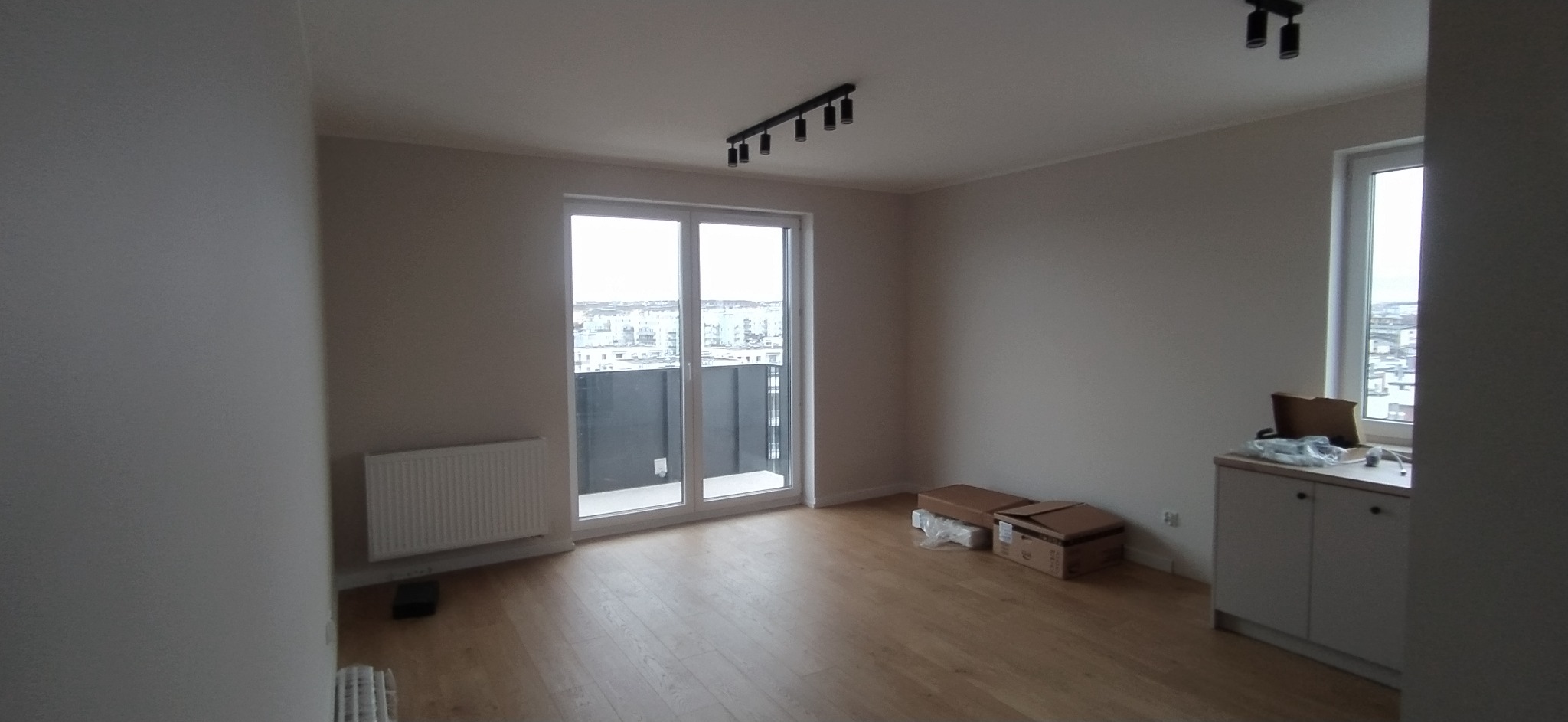 Pusty pokój z drewnianą podłogą, grzejnikiem, oknem balkonowym z widokiem na miasto, czarnym oświetleniem sufitowym i białymi szafkami w rogu.